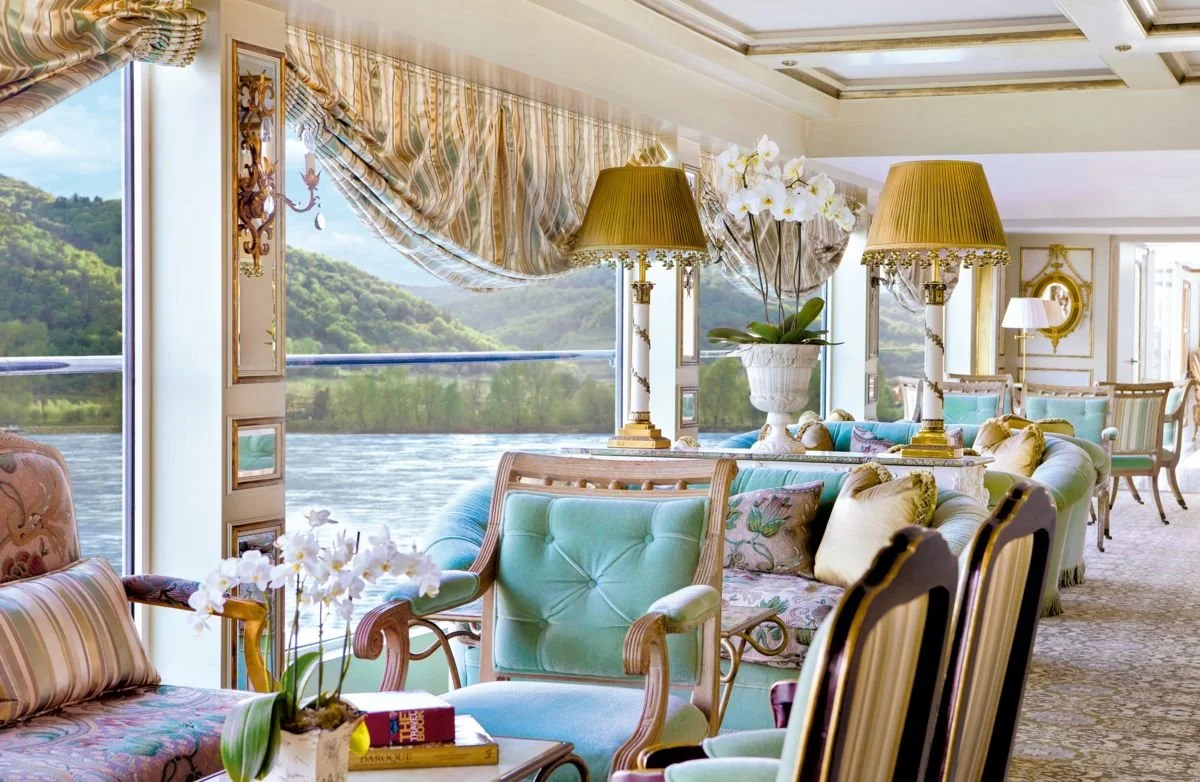 Luxurious sitting area with pastel furniture, decorative pillows, gold-accented lamps, large floral vases, and expansive windows overlooking a river and green hills.