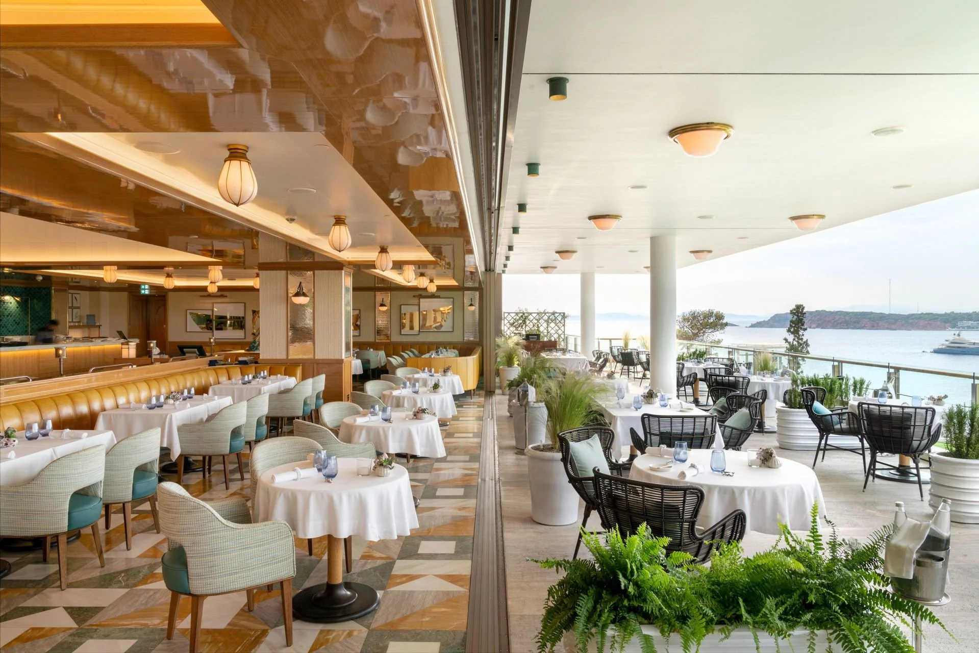 Elegant restaurant with tables and chairs set for dining, overlooking a body of water with boats and overlook view of a island or landmass in the background.