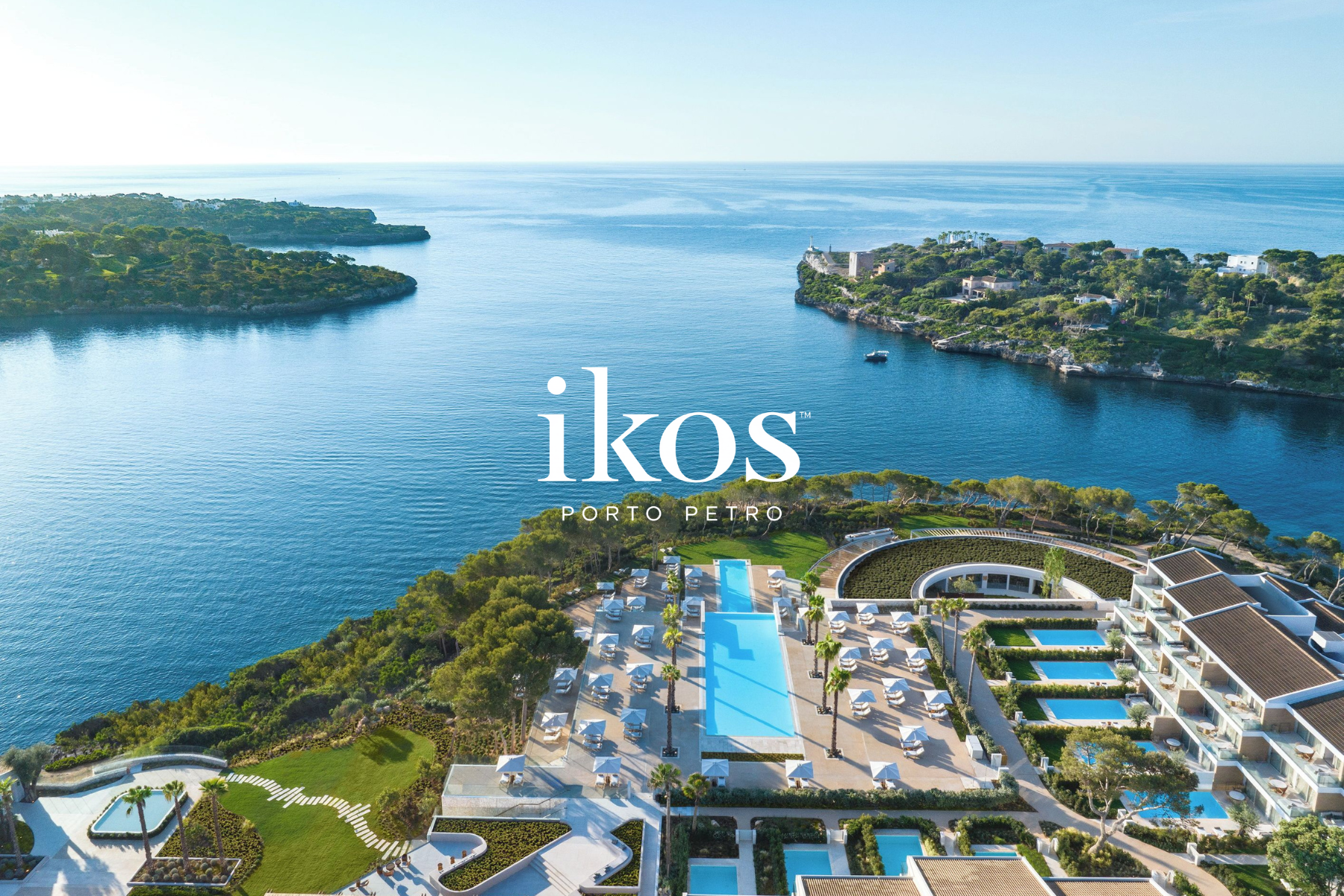 Aerial view of a luxury resort at Porto Petro, Ibiza, featuring multiple pools, sun loungers, palm trees, lush greenery, a coastline, and the ocean in the background.