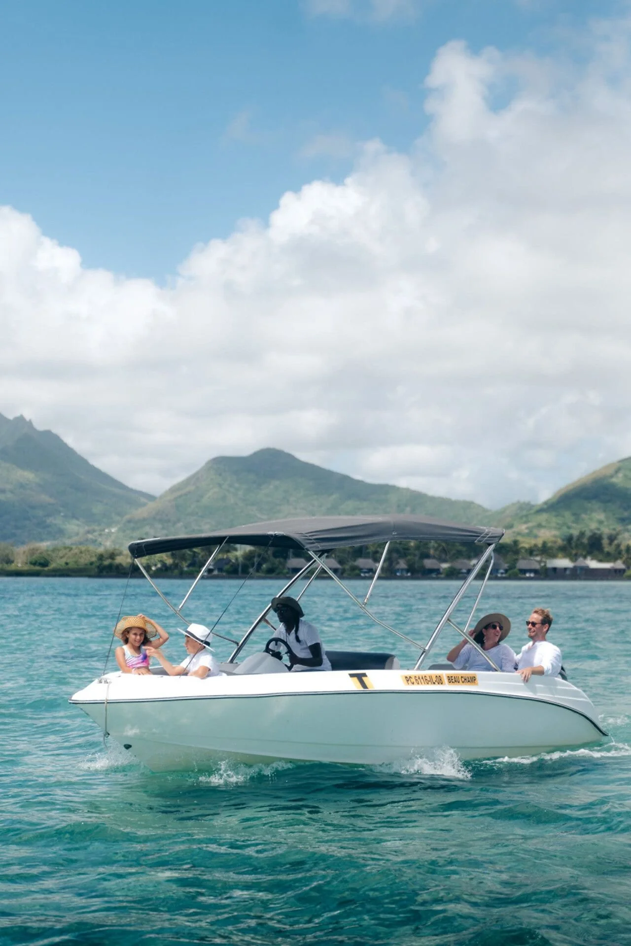 A group of five people on a white motorboat with a canopy, on a body of water with mountains and a cloudy sky in the background.