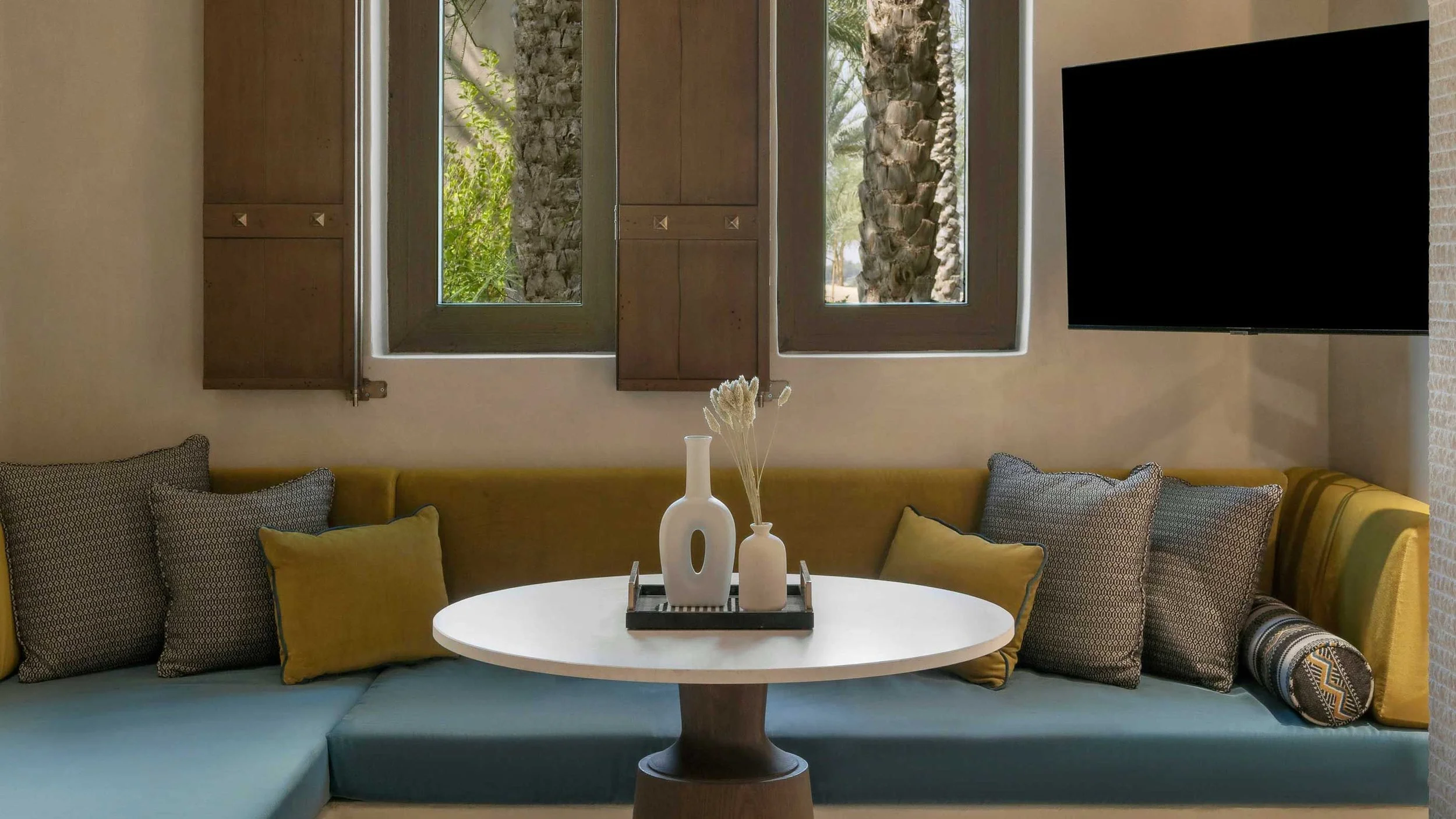 Living room with a yellow sectional sofa, multiple patterned pillows, a round white table with white vases and dried plants, three framed windows showing outdoor palm trees, and a large black flat-screen TV on the right wall.