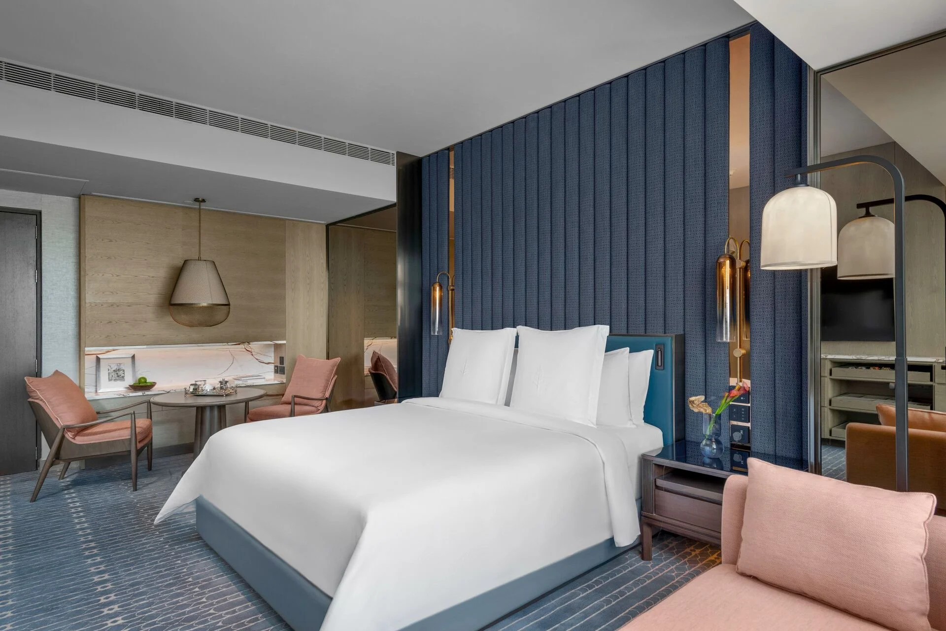 Modern hotel room with a large bed, blue upholstered headboard, and contemporary lighting fixtures. Small seating area with pink chairs and a round table in the background.