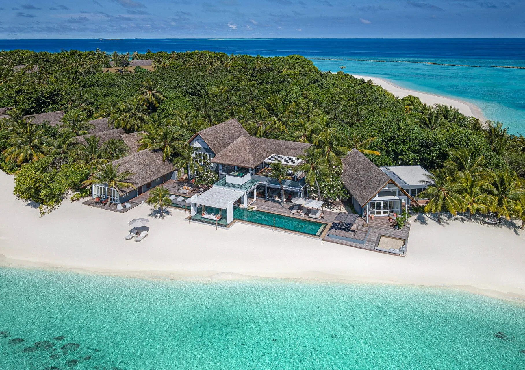 Luxury beachfront home with swimming pool surrounded by palm trees on white sandy beach, turquoise ocean water, and tropical greenery, with a clear blue sky overhead.