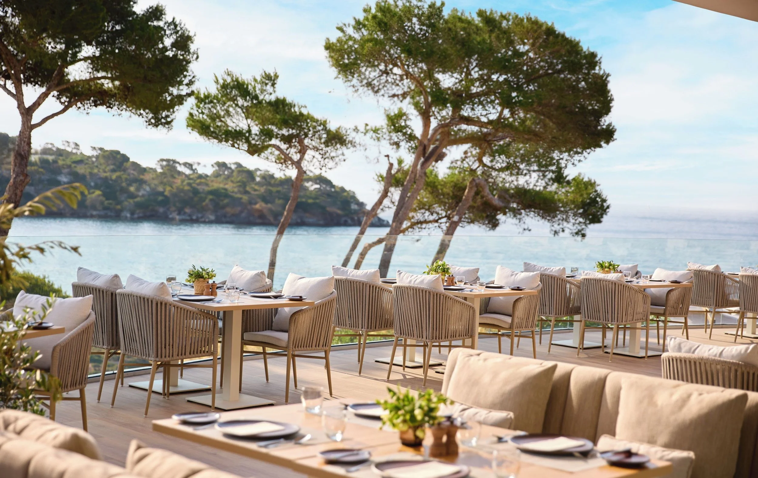 Outdoor restaurant with tables and chairs overlooking the ocean, with trees in the background.