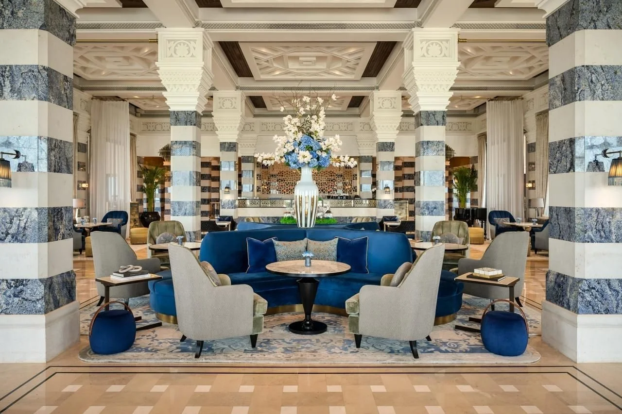 Elegant hotel lobby with marble columns, a blue velvet seating area with glass-top tables, and a large floral arrangement in a white vase at the center.