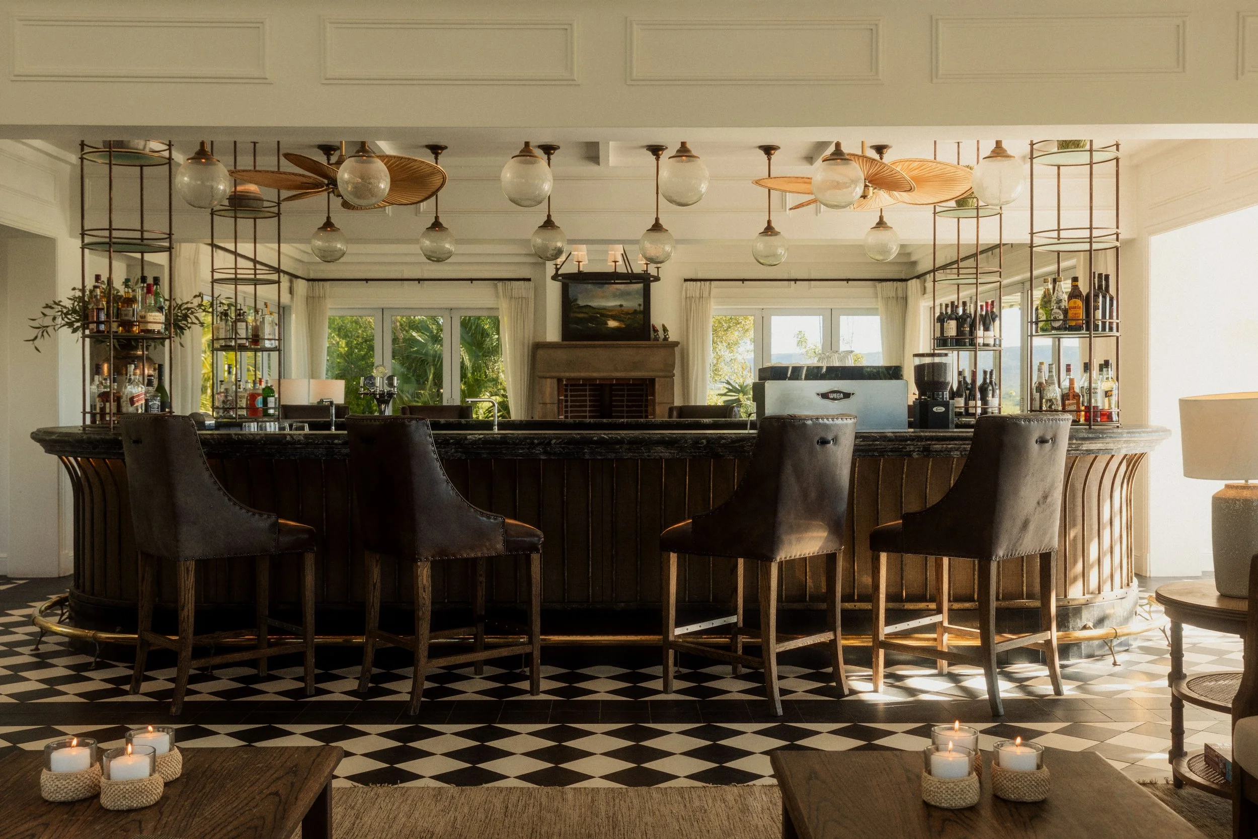 A stylish indoor bar with a marble counter, dark leather chairs, and a back bar decorated with bottles. The ceiling has globe and leaf-shaped light fixtures, and large windows with white curtains let in natural light. Candles are placed on coffee tables near the bar.