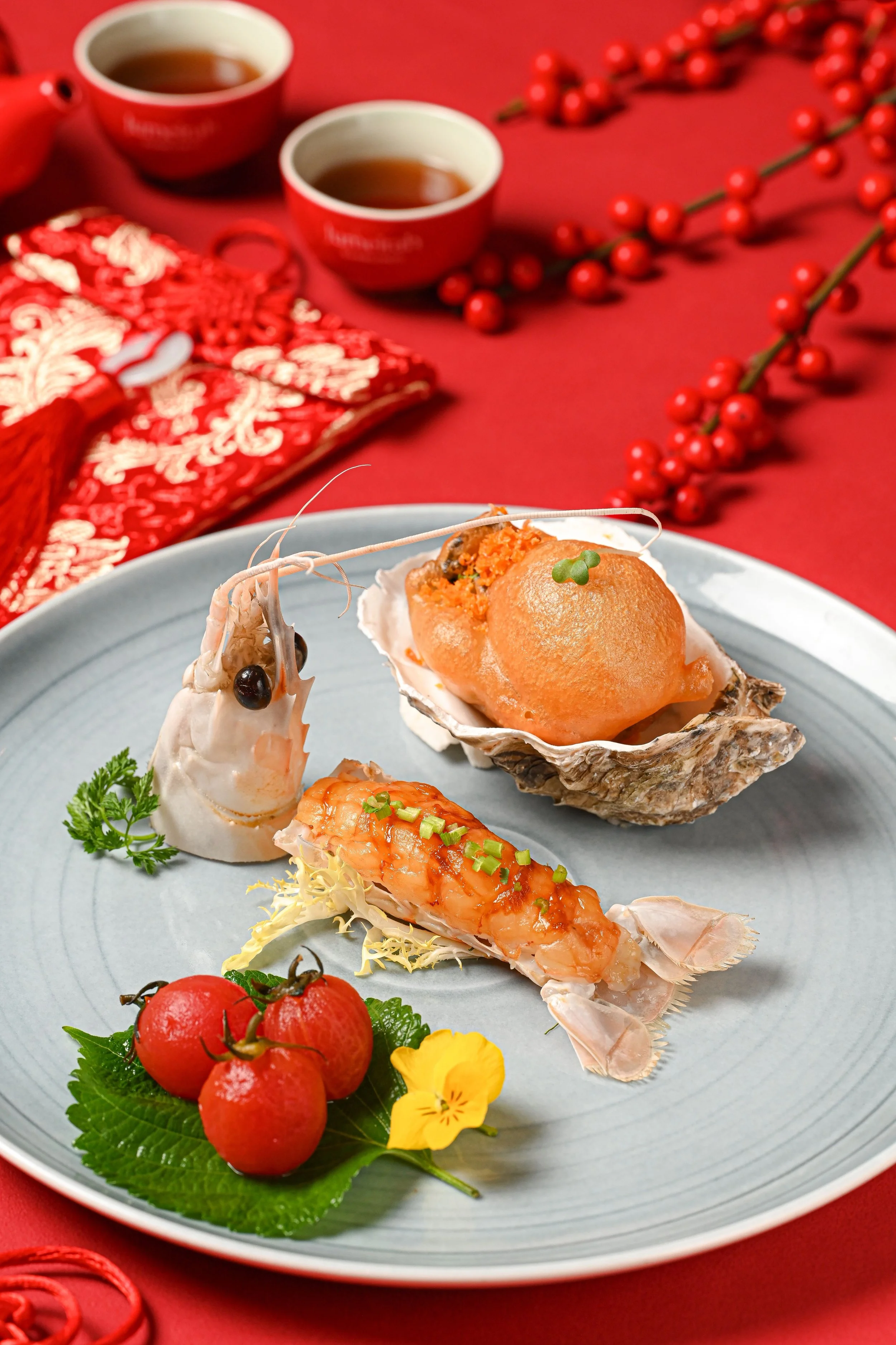 A blue plate with a variety of seafood including oysters, shrimp, and lobster, garnished with green herbs, small tomato, and yellow flower, placed on a red table with decorative red berries and a tea set in the background.