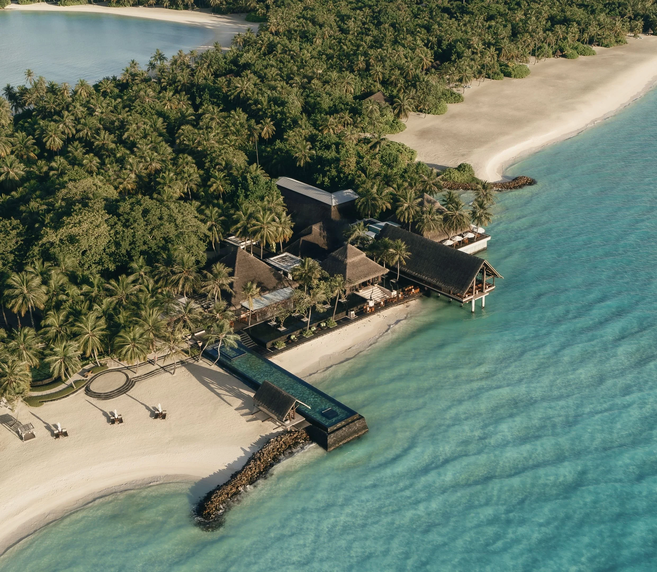 Aerial view of a tropical beach resort with sandy shoreline, turquoise water, lush green palm trees, and thatched-roof buildings.