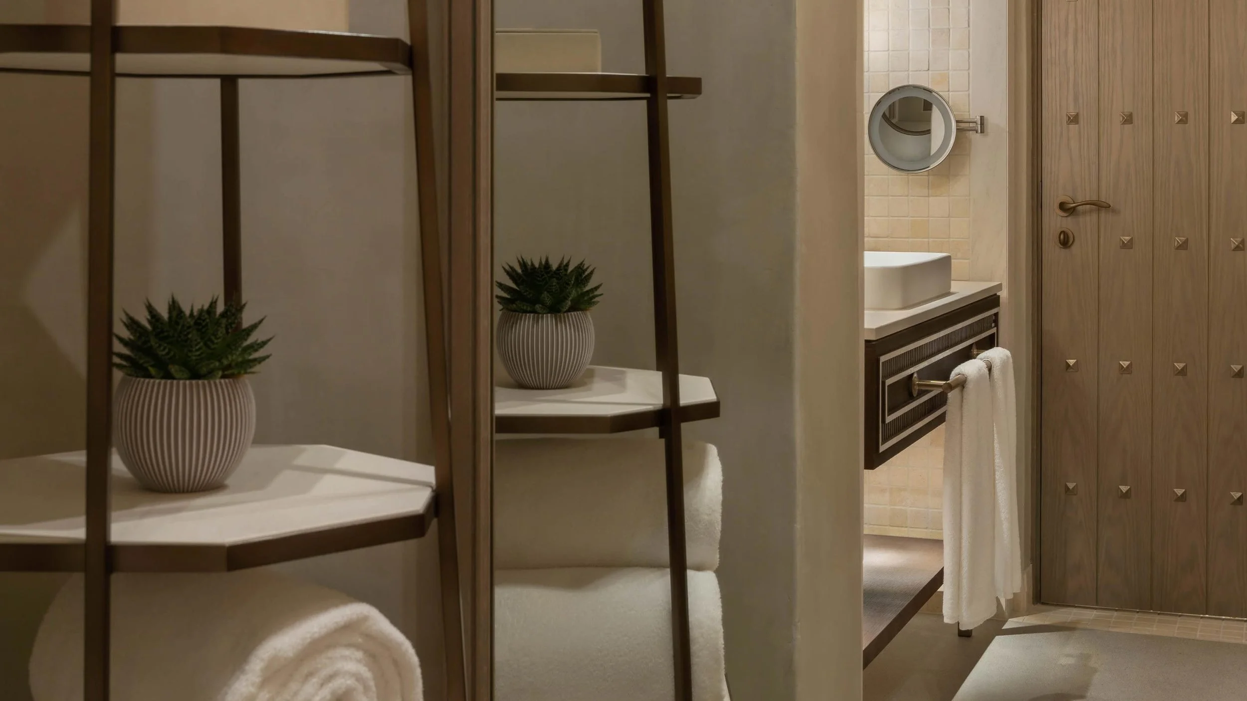 A bathroom with wooden accents, featuring a small white sink, a mirror, a towel rack with a white towel, a wooden door with geometric decorations, and a shelf with two small potted plants.