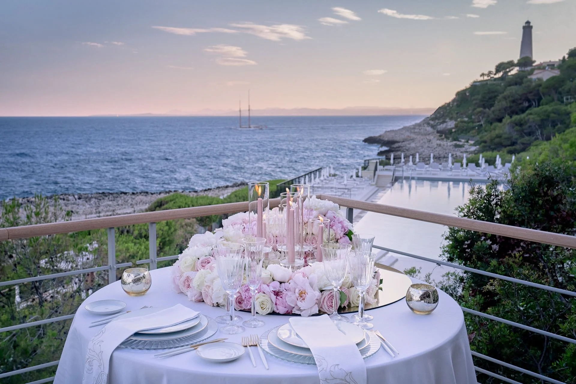 Elegant outdoor dining table decorated with pink candles and a floral centerpieces overlooking the ocean, with a sailboat in the distance, and a lighthouse on a hillside to the right.