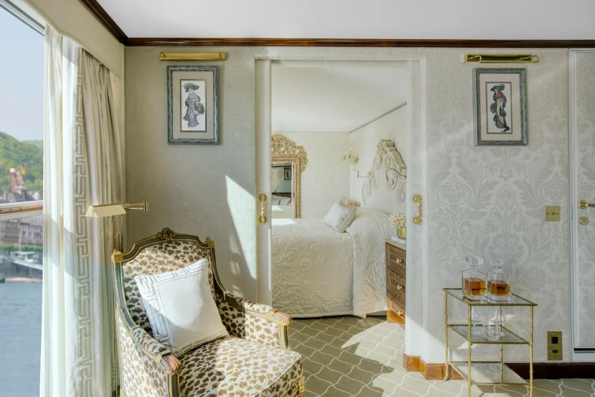 Luxury hotel room with a bed in the background, ornate furniture, and a window with curtains showing a river view.