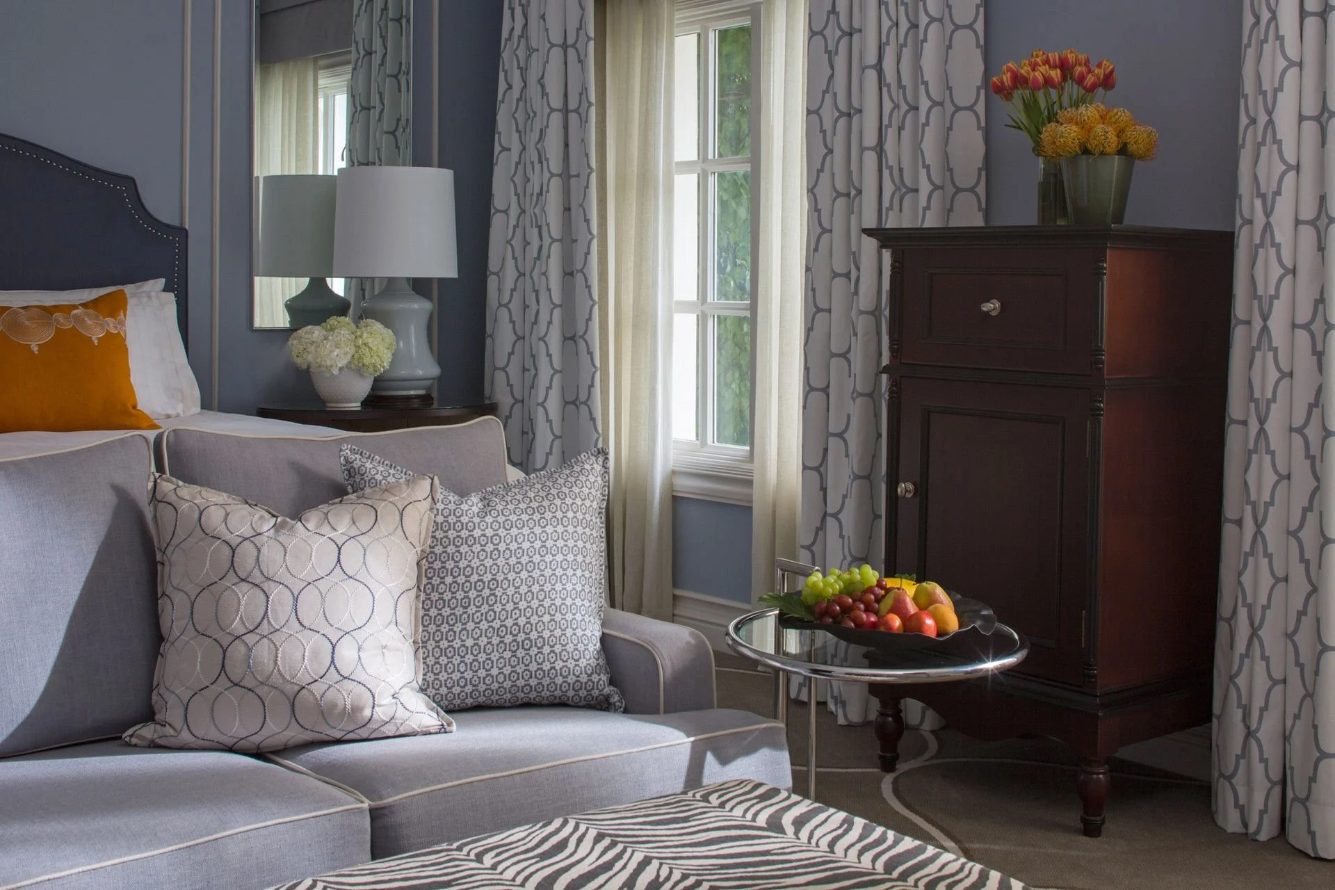 A cozy living room corner with a light gray couch adorned with patterned throw pillows, beside a dark wood side table with a white lamp and flowers, a window with patterned curtains, and a dark wood cabinet with a large vase of orange and yellow flowers. A glass-topped side table holds assorted fruits.