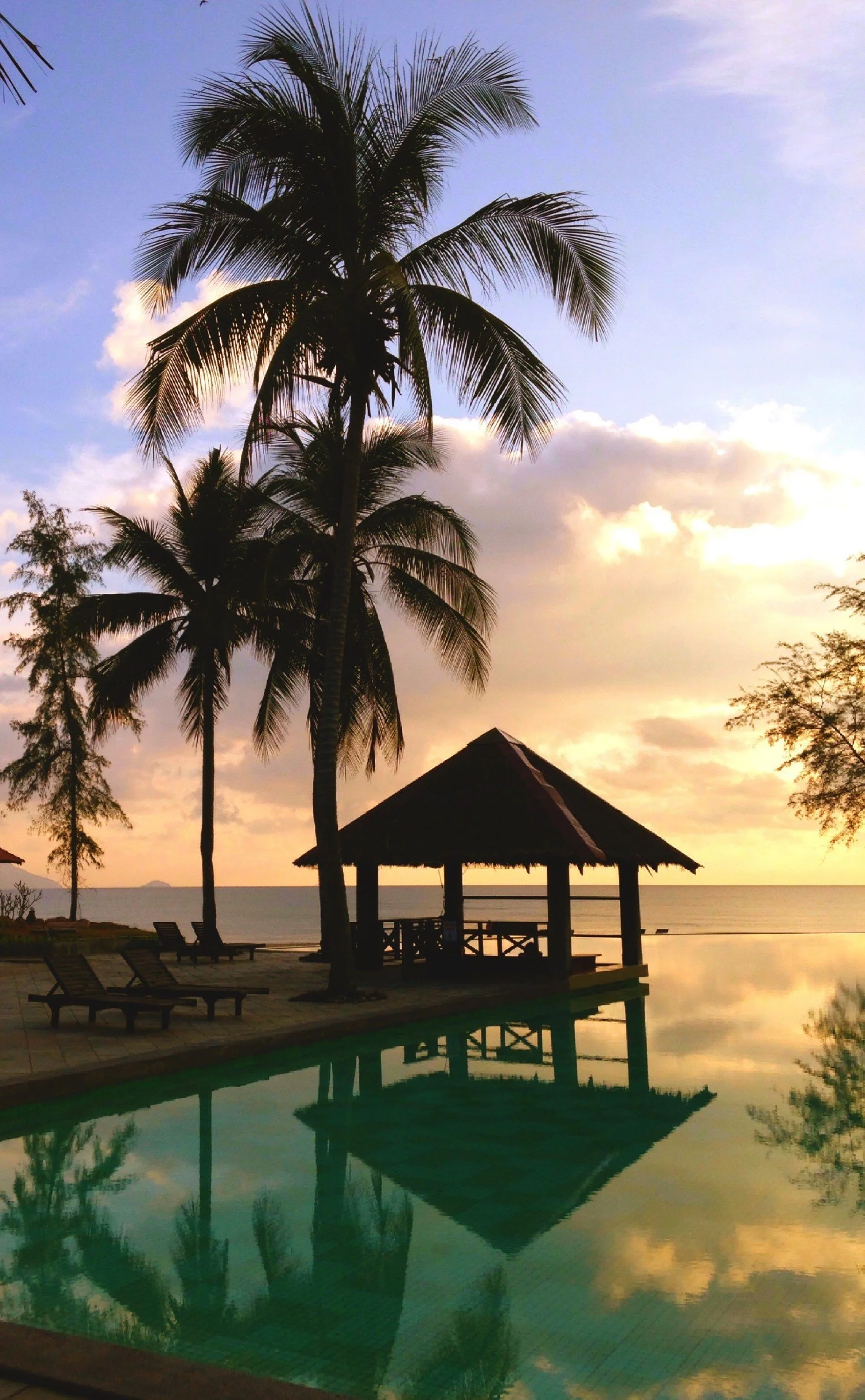 Sunset over a tropical beach with palm trees, a pool, lounge chairs, and a thatched roof hut reflecting in the water.