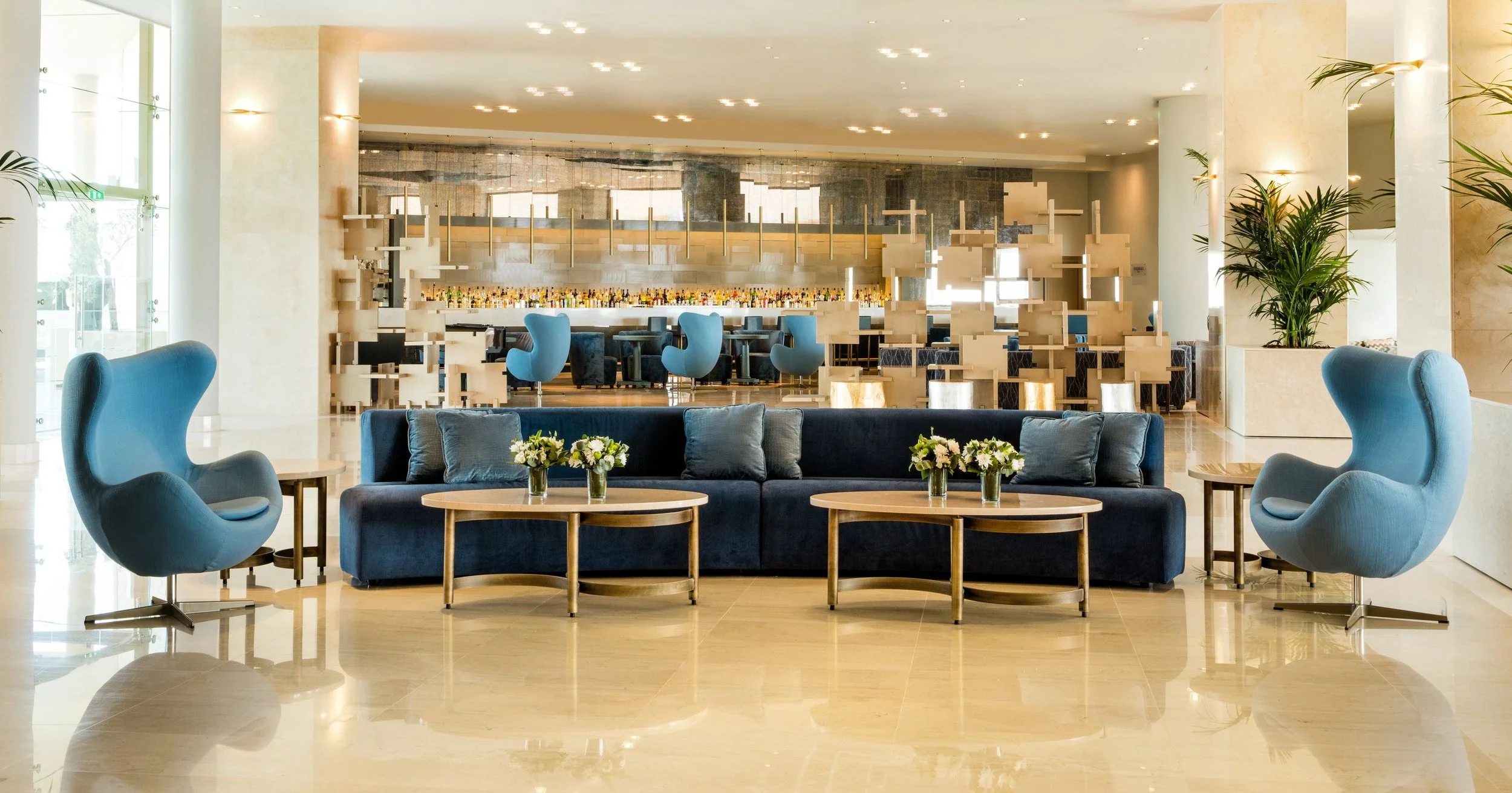 Luxurious hotel lobby with modern design, featuring a blue sofa and armchairs, decorative coffee tables with flower arrangements, large windows, and a bar area in the background.
