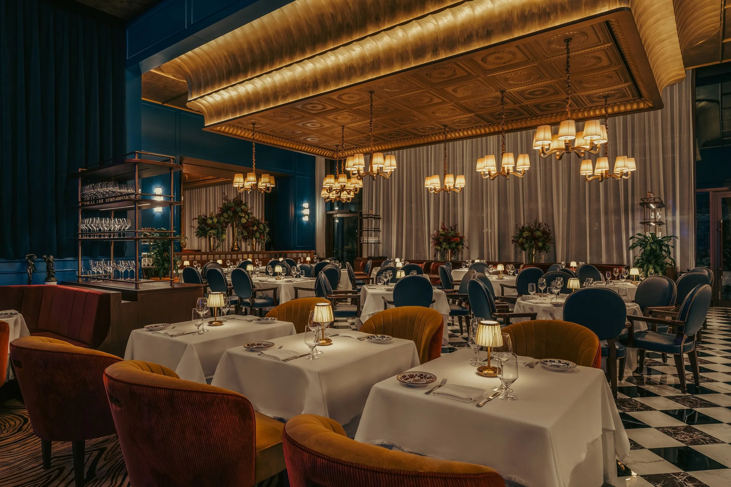 Interior of a luxurious restaurant with white tablecloths, colorful velvet chairs, elegant chandeliers, floral centerpieces, and checkered black and white flooring.