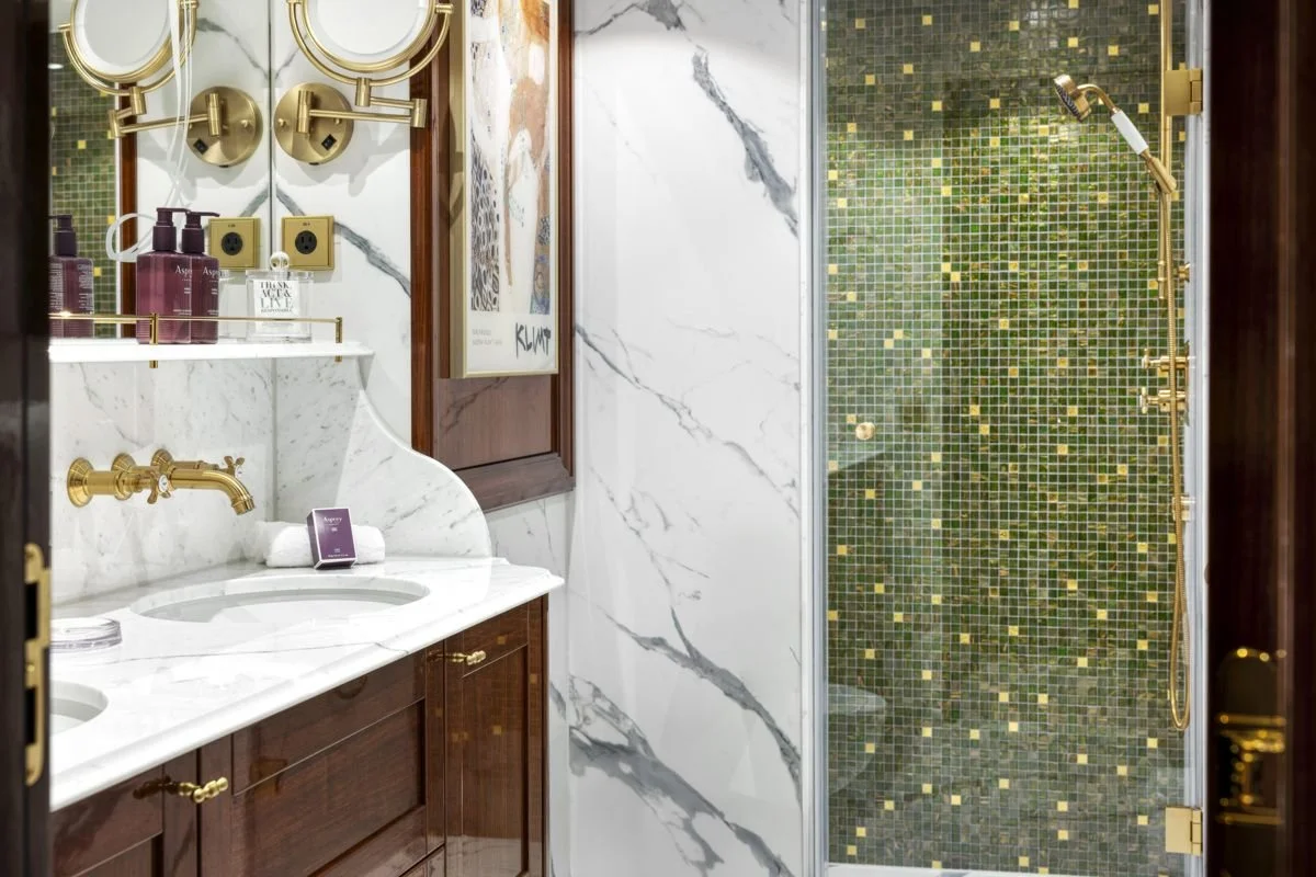 A luxurious bathroom with white marble countertops, a double sink, gold fixtures, a wall-mounted mirror, a framed piece of art, and a green mosaic-tiled shower with gold fixtures.