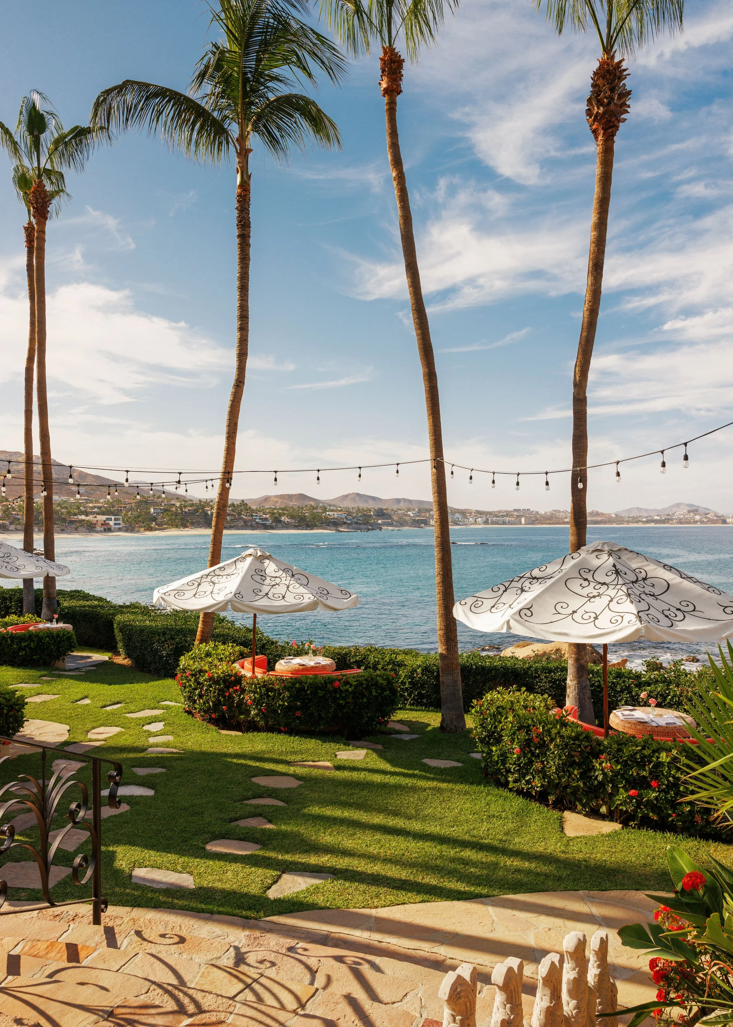 View of a coastal scene with palm trees, a fence, umbrellas, and a harbor in the background during the daytime.