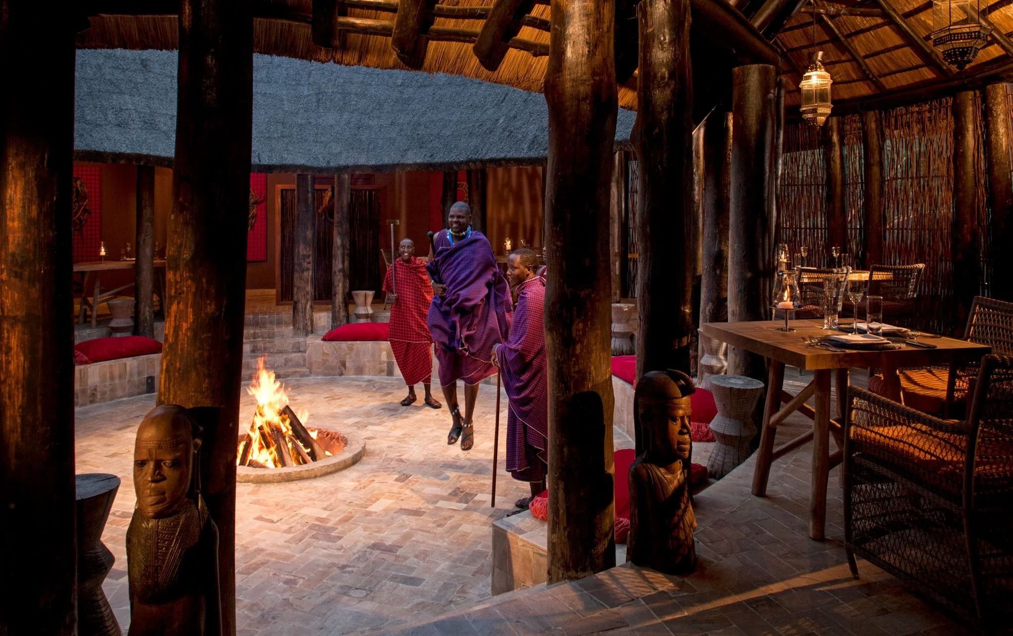 People in traditional Maasai attire standing around a campfire inside a thatched-roof structure with wooden support beams, masks, and decorated tables.