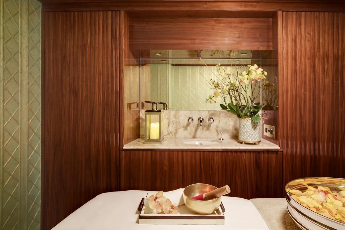 A cozy indoor space featuring a wooden privacy divider with a mirror, a marble countertop with a vase of orchids, a lantern with a candle, and a tray with flowers and a pestle. A bowl of floating flower petals is visible on the right side.