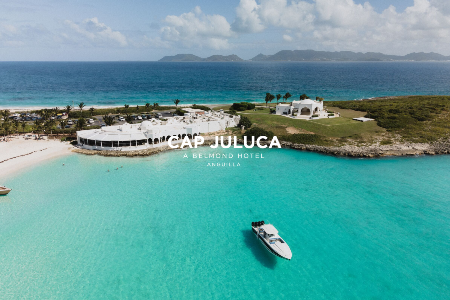 Aerial view of Cap Juluca resort on Anguilla, featuring white buildings, palm trees, clear turquoise water, a sandy beach, cars parked near the resort, and a boat moving through the water with mountains in the background.