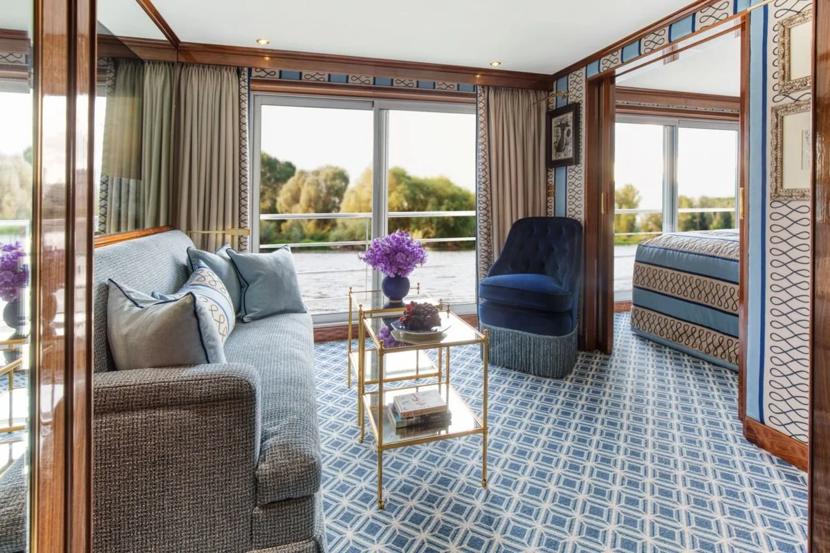 Living room with large window overlooking water, blue and beige patterned decor, blue couch with pillows, dark blue armchair, glass coffee table with flowers and books.