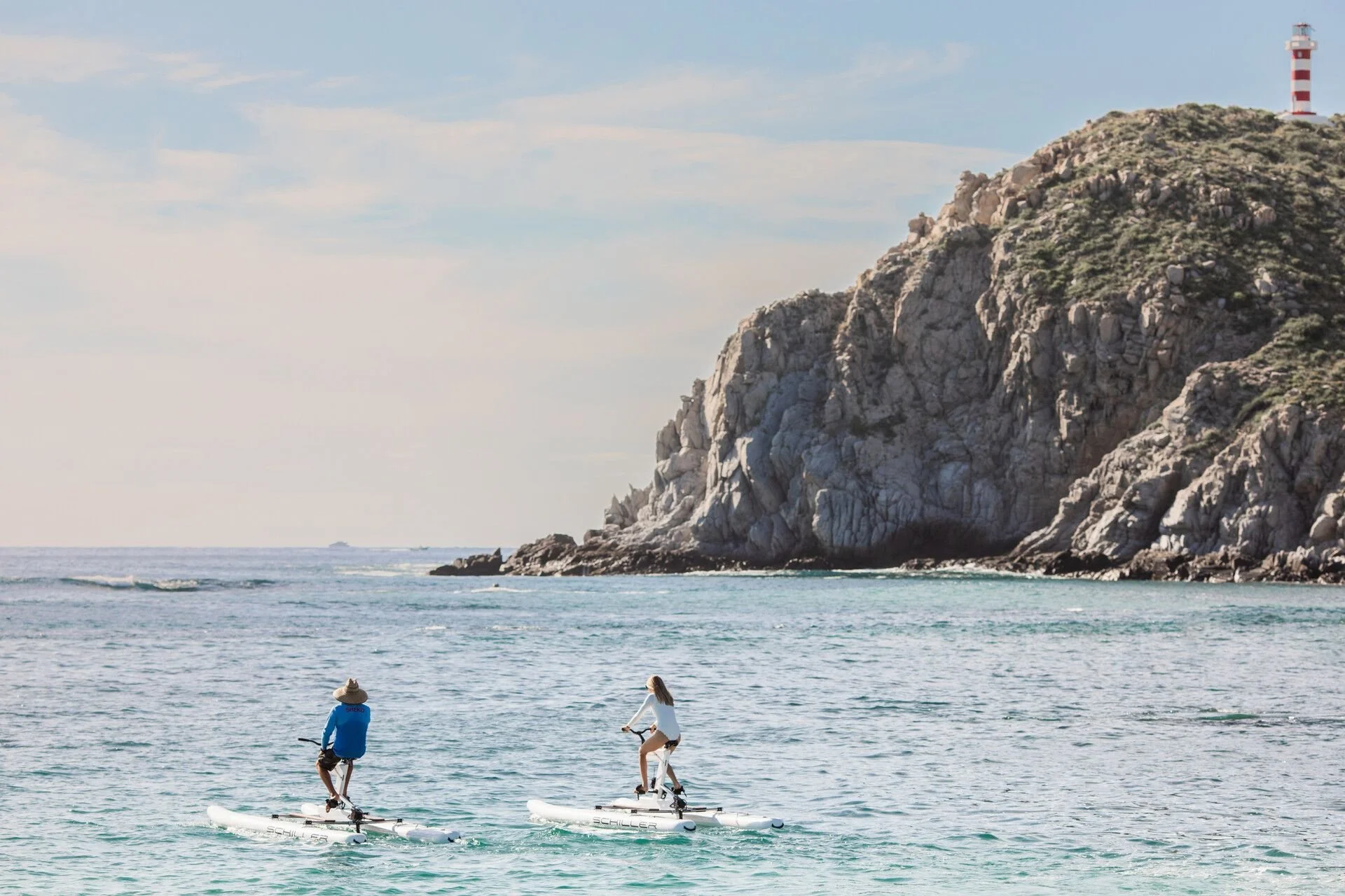Two people riding paddleboards on the ocean near a rocky cliff with a lighthouse on top.