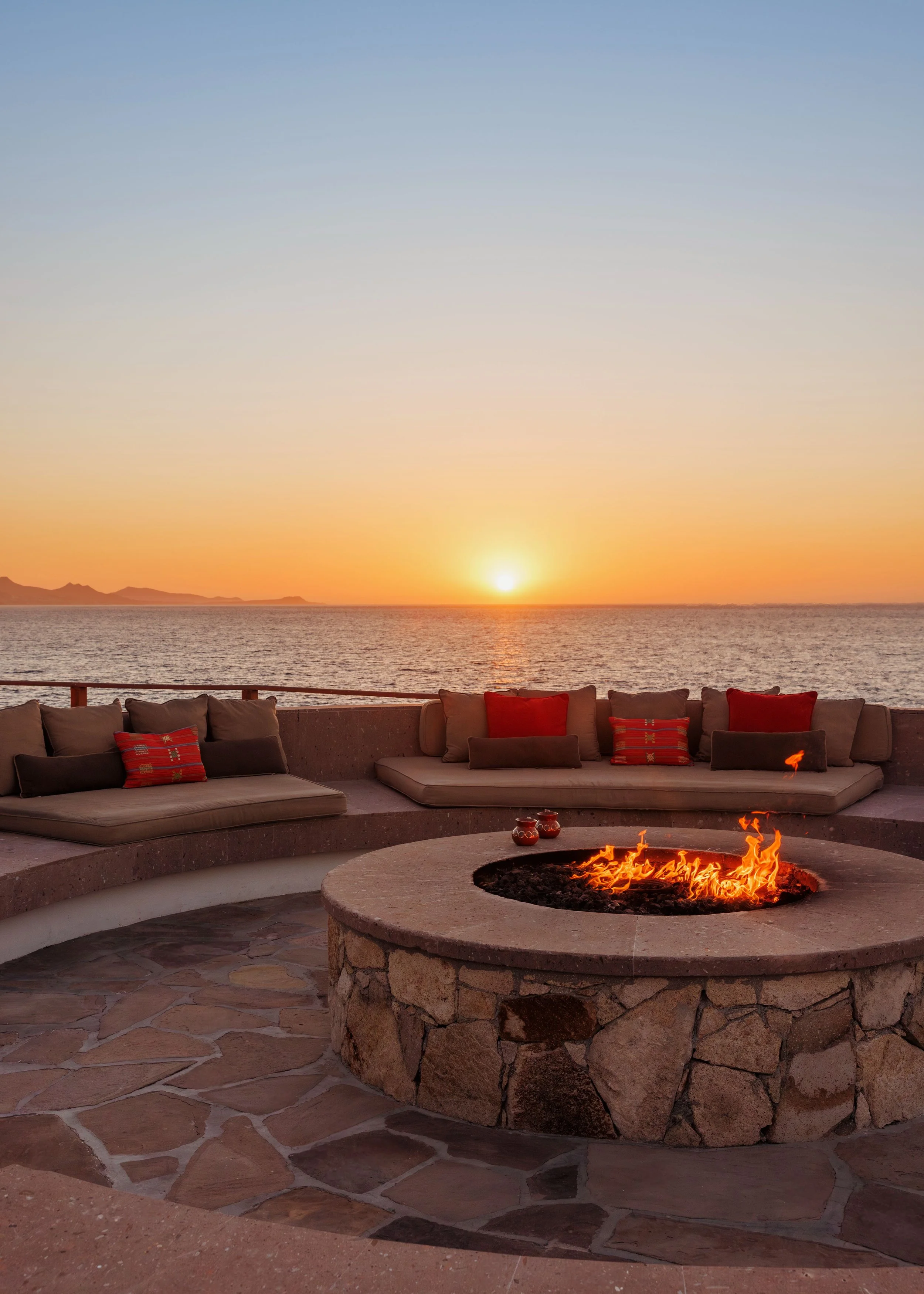 Outdoor seating area with cushioned benches and pillows facing a fire pit, overlooking the ocean during sunset.