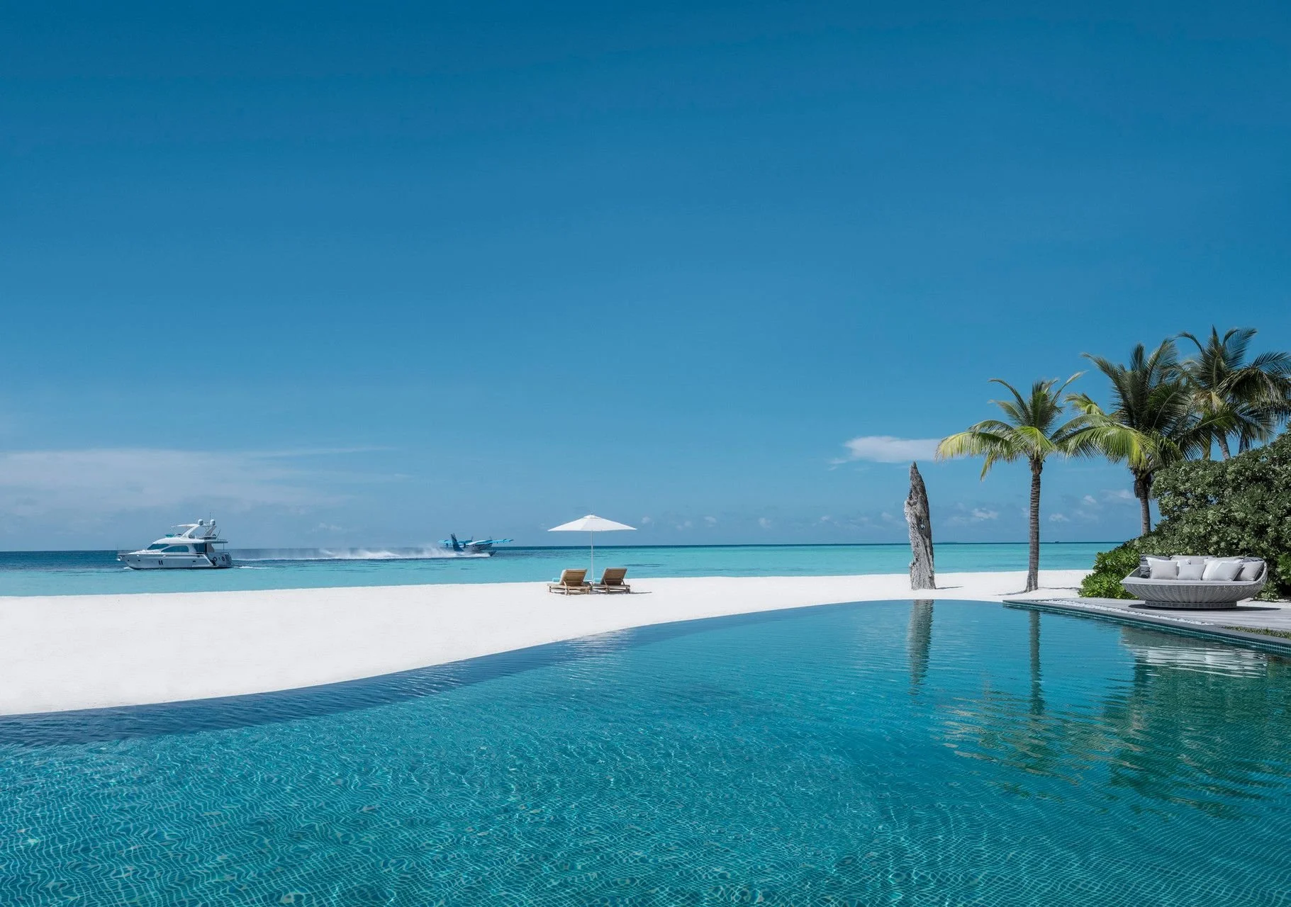 Luxurious beach scene with an infinity pool, white sand, palm trees, lounge chairs with an umbrella, a boat and jet skis on the water, and a clear blue sky.