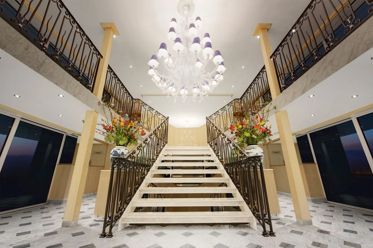 Inside a luxurious home featuring a double staircase with ornate black railings, large flower arrangements in ceramic vases on either side, a grand chandelier hanging from the ceiling, marble flooring, and large windows showing an outdoor sunset view.