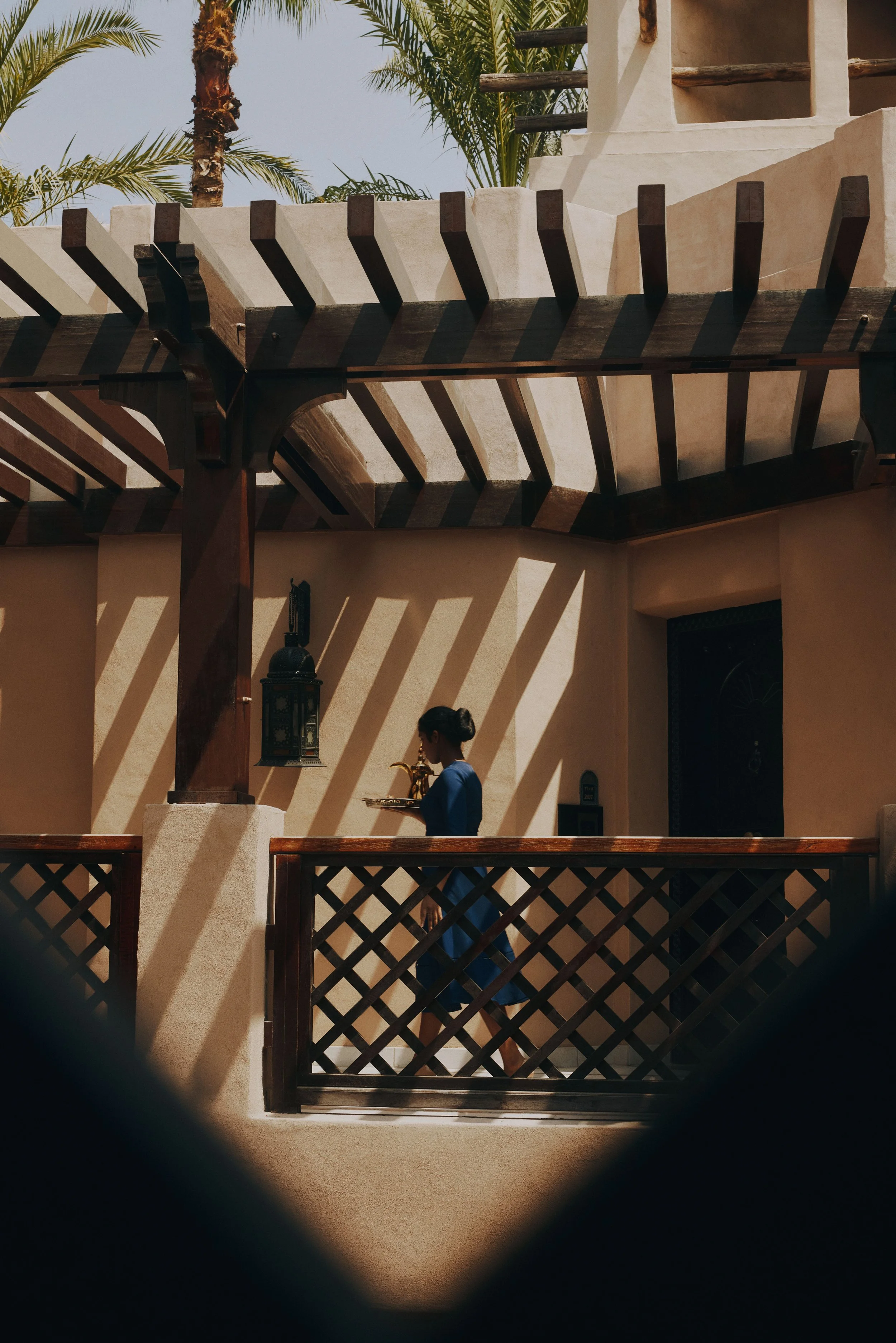 A woman in traditional attire holding a tray of teas or similar drinks, standing on a balcony with a lattice railing. The scene is cast in warm sunlight with palm trees in the background, and architectural elements with wooden beams and lanterns are visible.