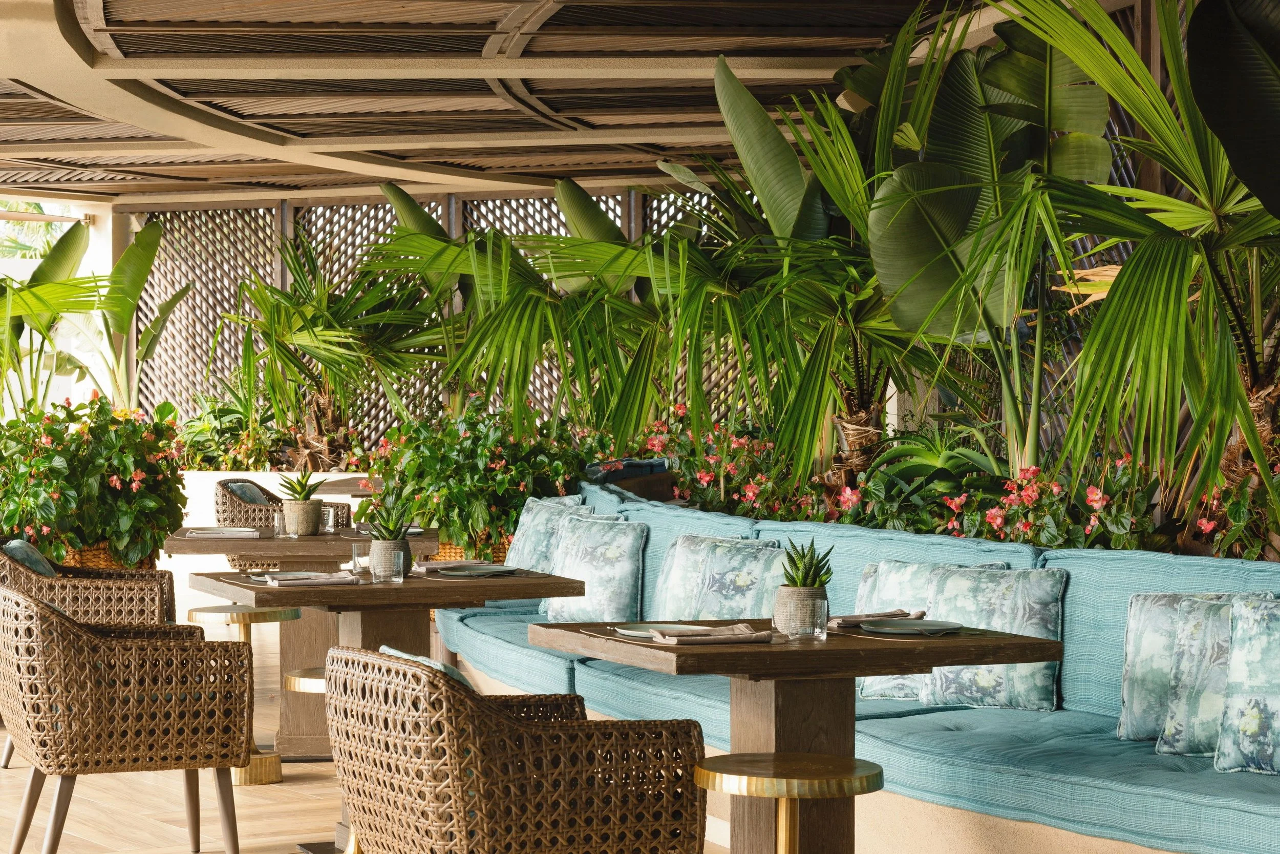 Outdoor patio with wicker chairs, wooden tables, floral and green potted plants, and a cushioned seating area with blue pillows. A wooden lattice privacy screen separates the space.