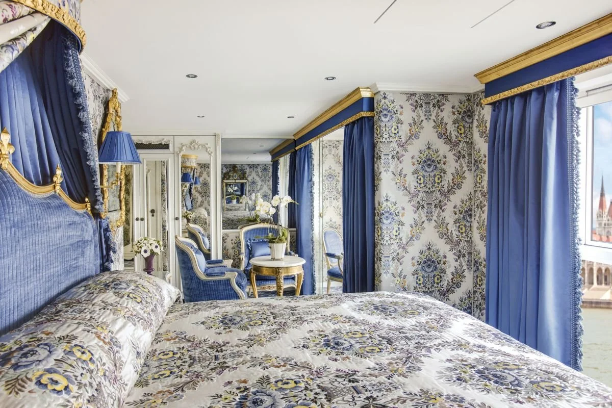 Luxurious bedroom with blue and gold accents, floral patterned wallpaper, and matching curtains and bedding, featuring a mirrored wardrobe, small seating area, and a window with a view of a historic building.
