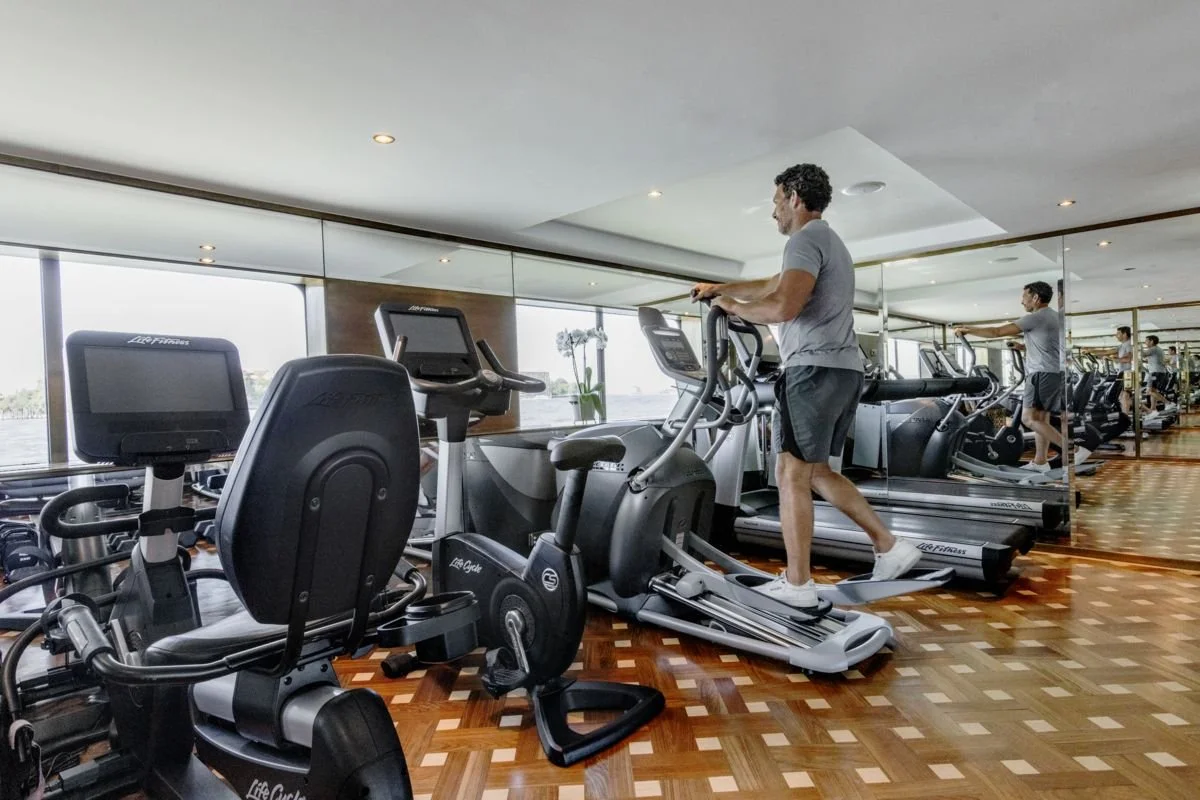 A man running on a treadmill in a gym with large mirror and windows.
