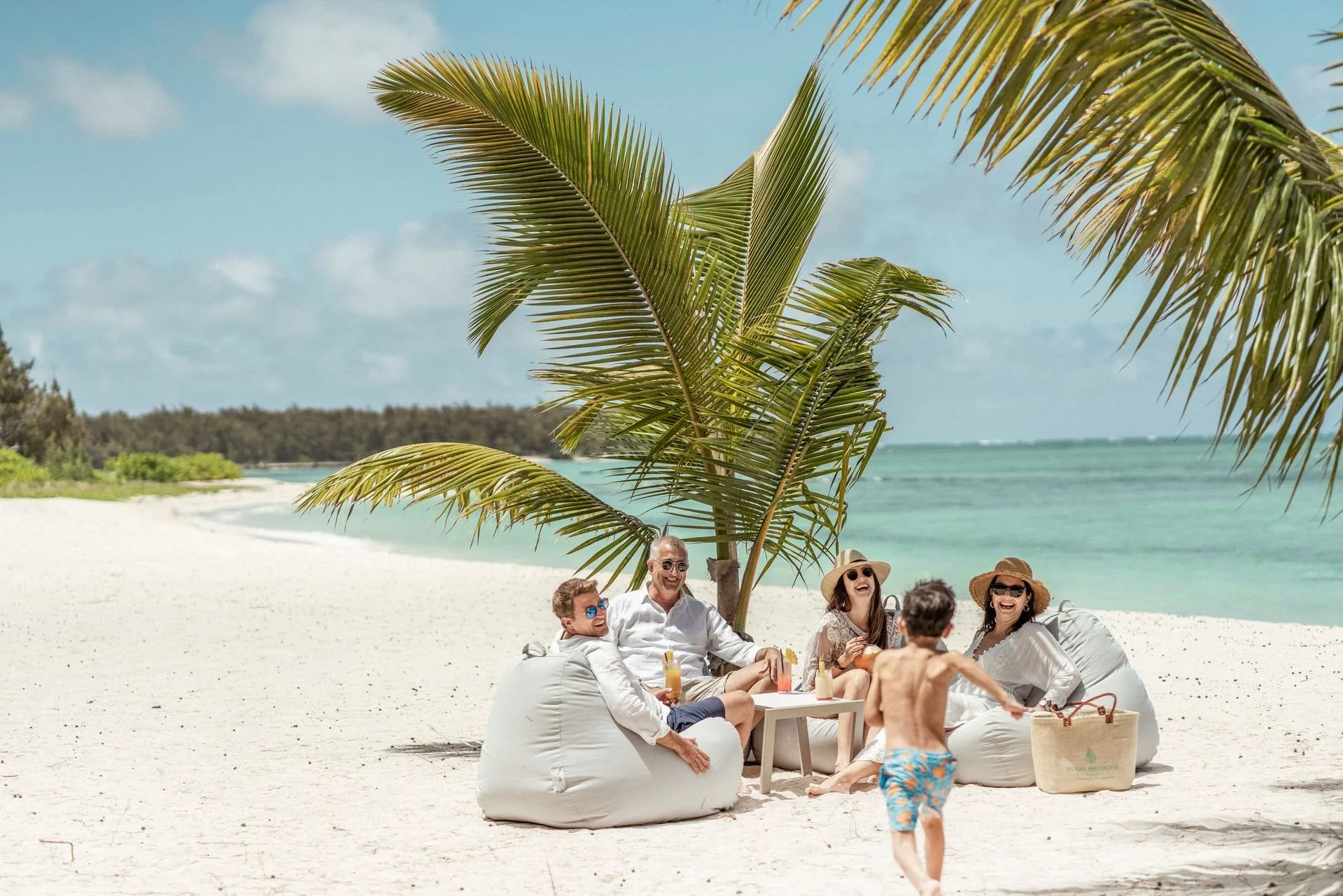 A group of people enjoying a sunny day on the beach, sitting on bean bag chairs under a palm tree, with drinks on a small table, and smiling while socializing.