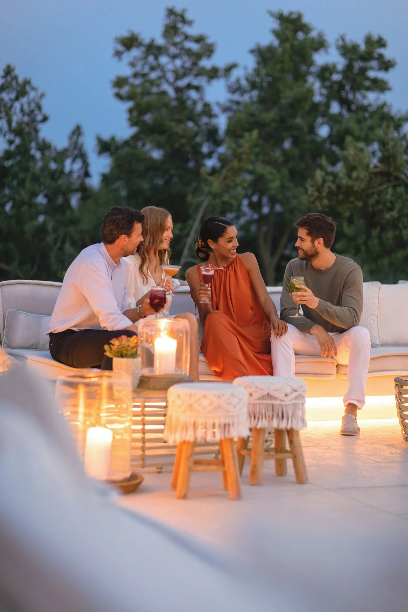 Four people sitting on a white outdoor sofa during evening, enjoying drinks and conversation, with candles and small tables nearby, in a setting with trees in the background.