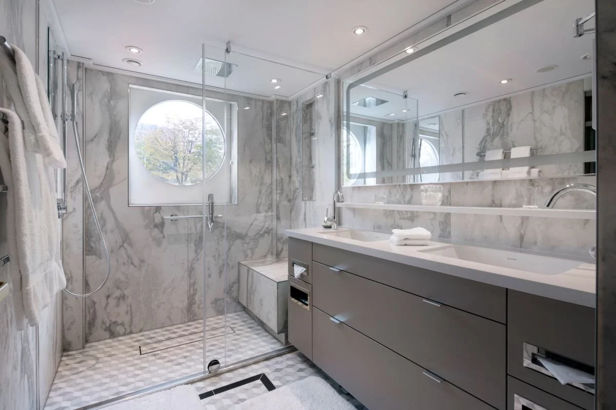Bathroom with a marble shower area, a large mirror above a gray vanity with a countertop, a round window letting in natural light, and white towels on the counter.