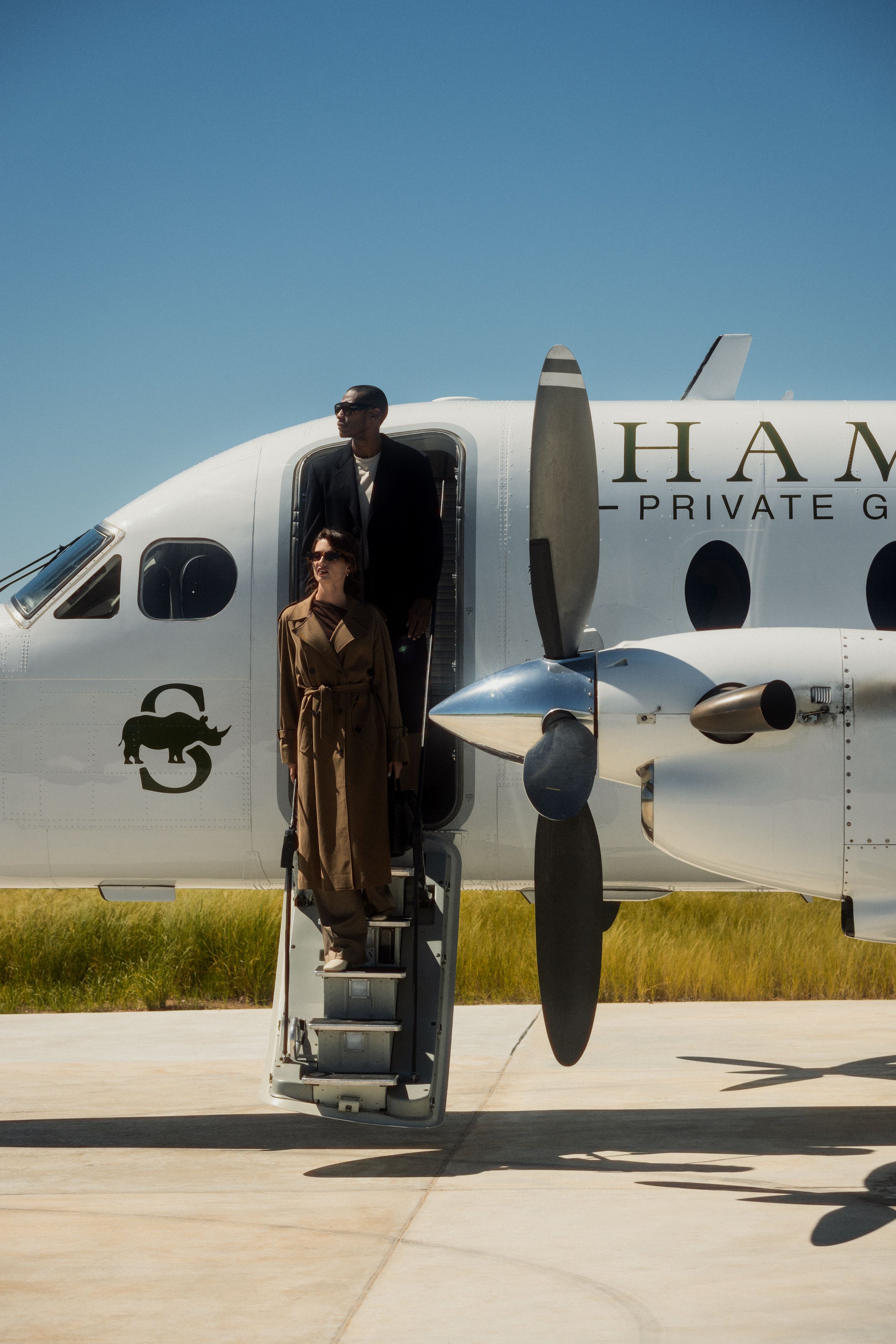Two people descending the stairs of a private jet on a sunny day, with a clear blue sky and grassy field in the background. The jet displays a logo with a black panther and partially visible text.