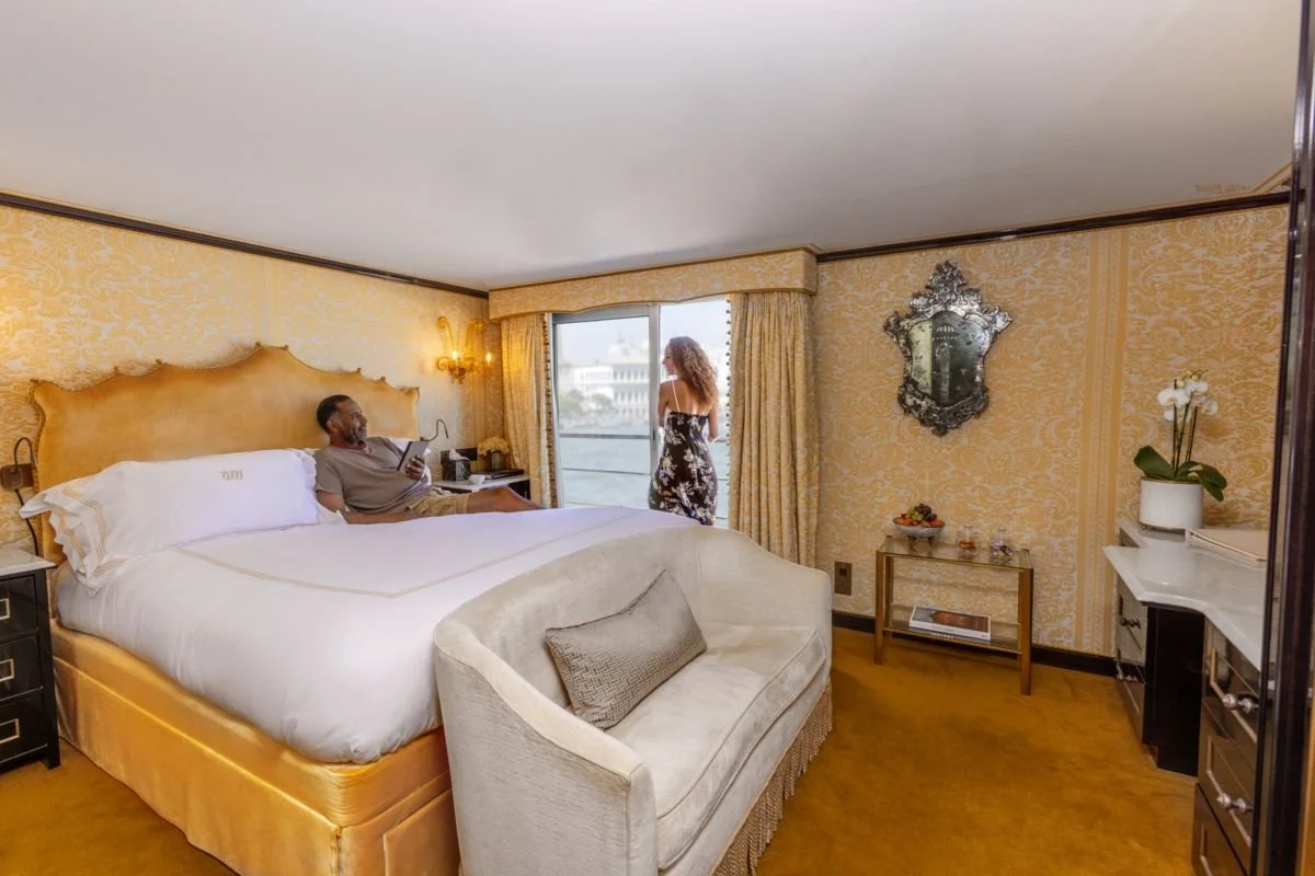 A hotel room with a large bed, a man sitting on the bed using a phone, a woman standing by the window looking outside, and elegant decor including a mirror and floral wallpaper.