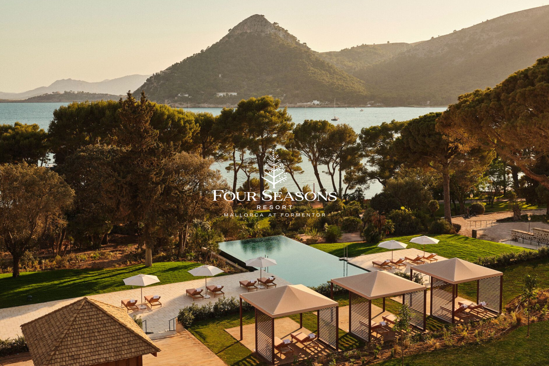 View of the Four Seasons Resort in Mallorca at Formentor, showing a swimming pool area with lounge chairs and umbrellas, surrounded by lush green lawns and trees, with mountains and water in the background.