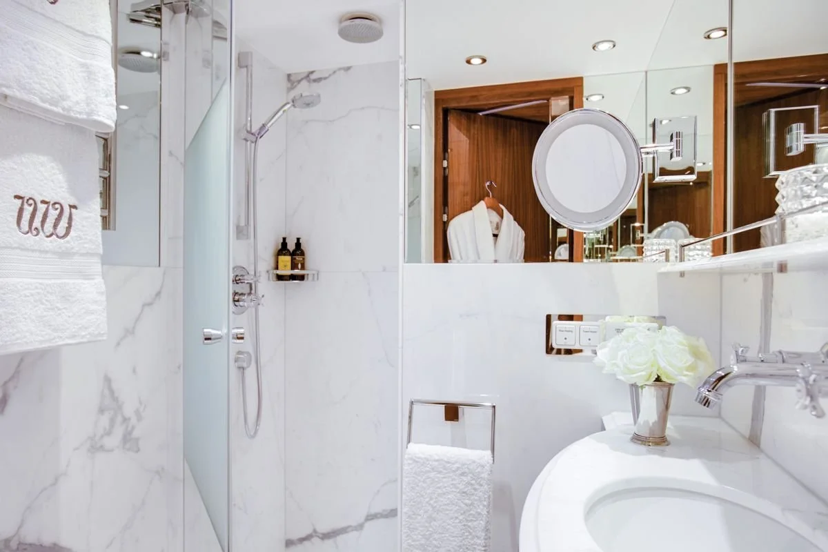 A luxurious bathroom with marble walls, a walk-in shower with glass door, a towel rack with a white towel, a vanity with a sink and flowers, a mirror, and a robe hanging on a wooden door.