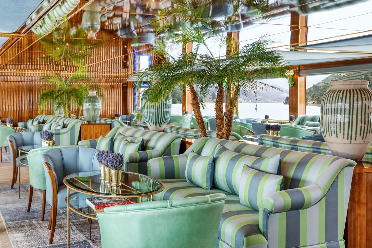 Interior of a bright, airy lounge or restaurant with striped green and blue furniture, large vases, lush palm trees, and a view of water outside.