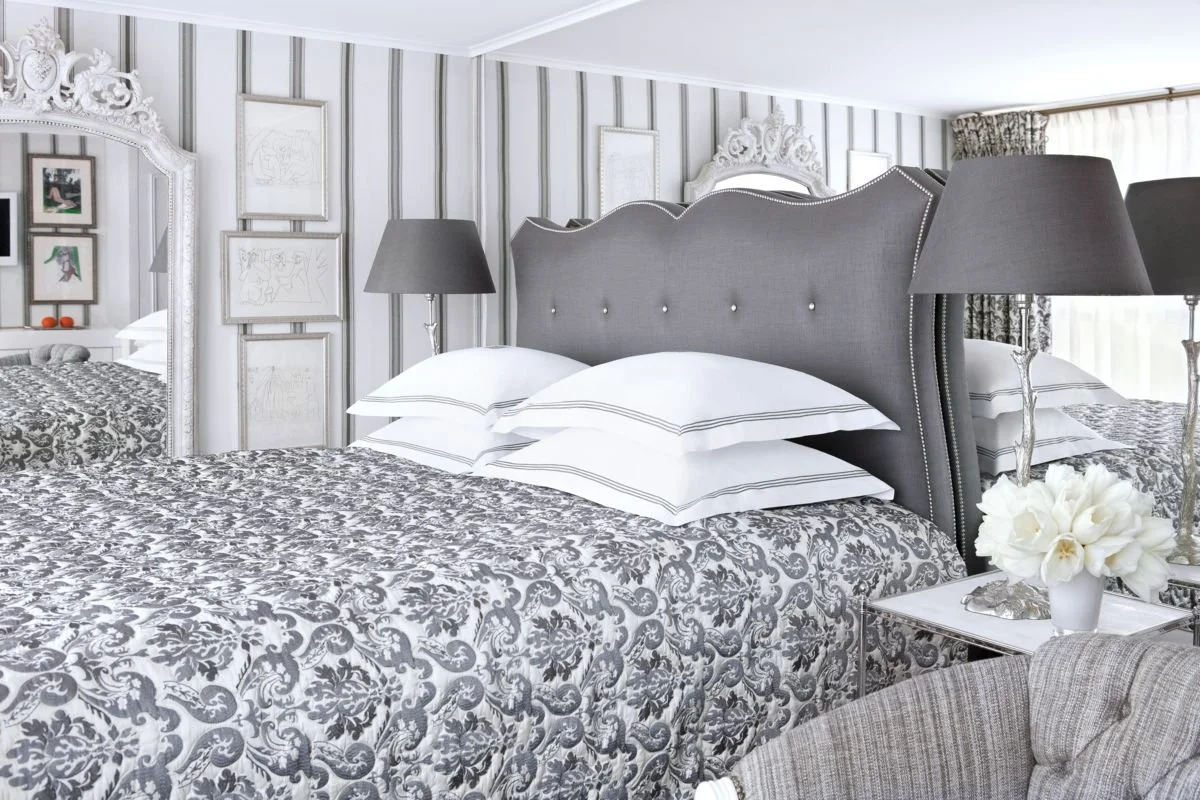 Elegant bedroom with a large bed featuring a gray upholstered headboard, white bedding, and multiple pillows, flanked by matching gray lamps, with decorative framed art on the striped wallpaper wall and a large mirror.