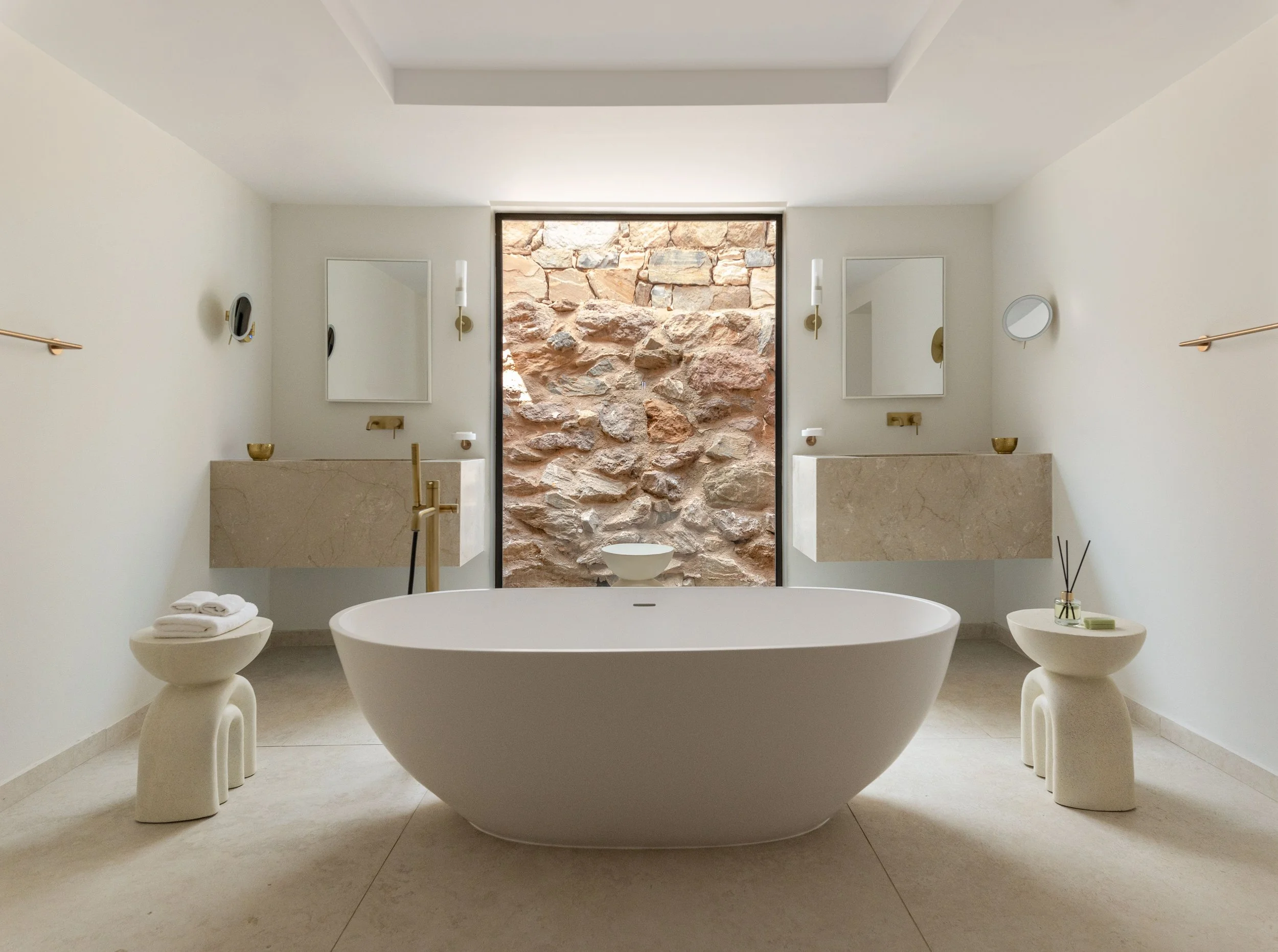 A modern bathroom with a freestanding bathtub, two side chairs, a stone wall with a large window, and minimal decor.