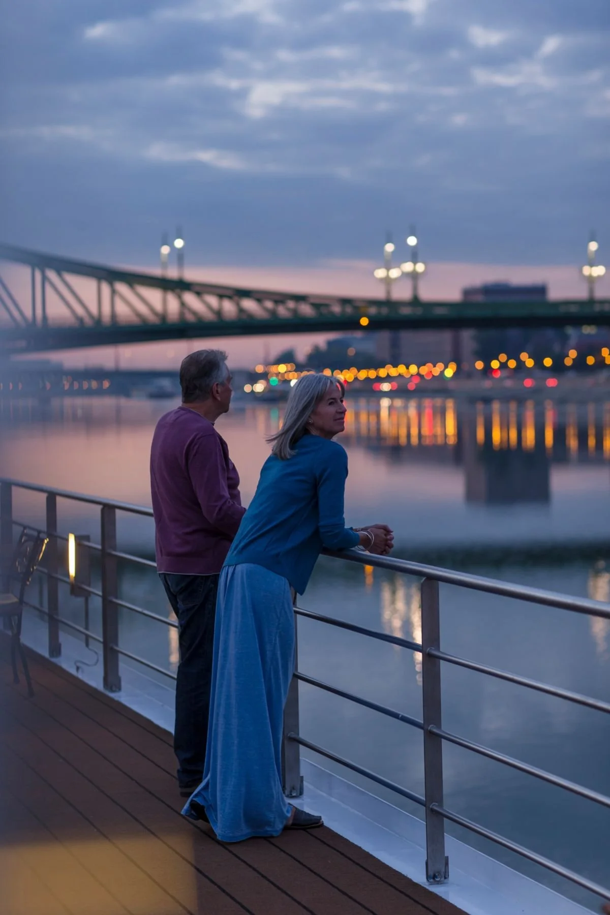 A man and woman stand on a boat deck by the water at dusk, gazing at a bridge with city lights reflecting on the water.