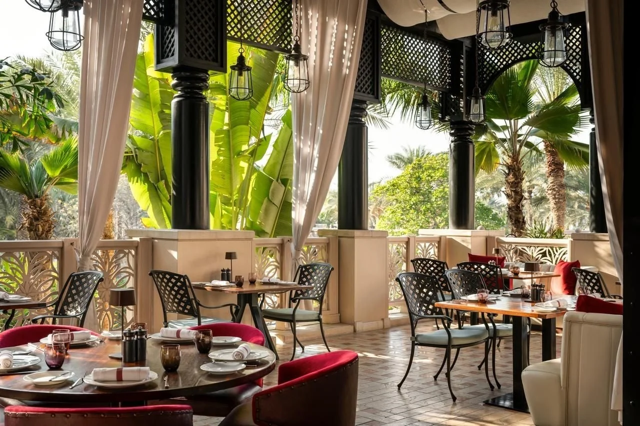 An outdoor restaurant patio with tables set for dining, surrounded by lush green tropical plants and tall palm trees, with black columns, decorative curtains, and hanging lantern-style lights.