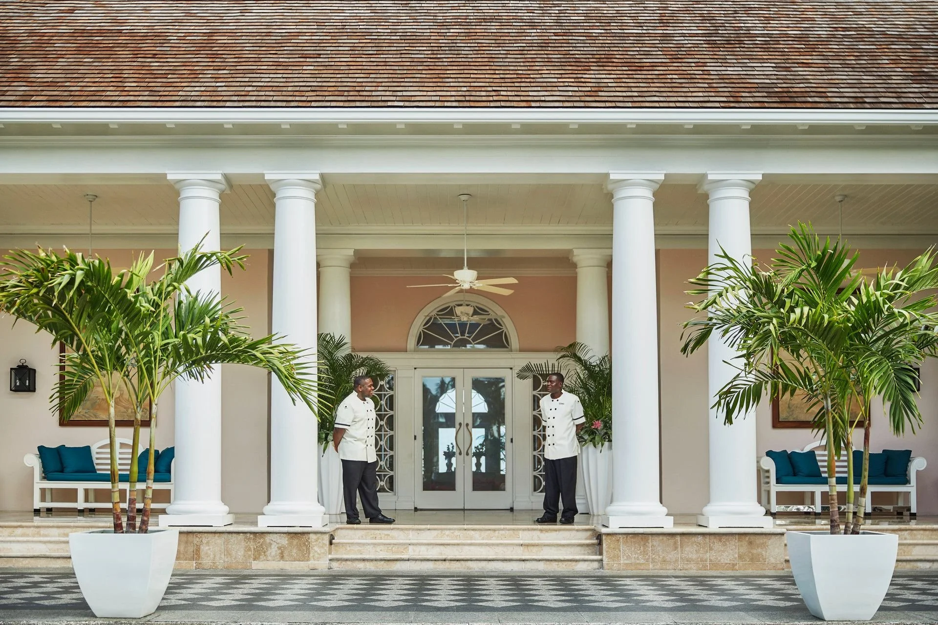 Entrance of a hotel or restaurant with two staff members standing under a portico with white columns, decorative plants, and seating area.