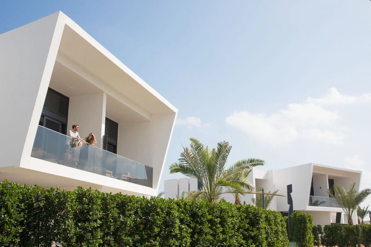 Modern white buildings with large glass windowed balconies, surrounded by green hedges and palm trees, under a partly cloudy sky.