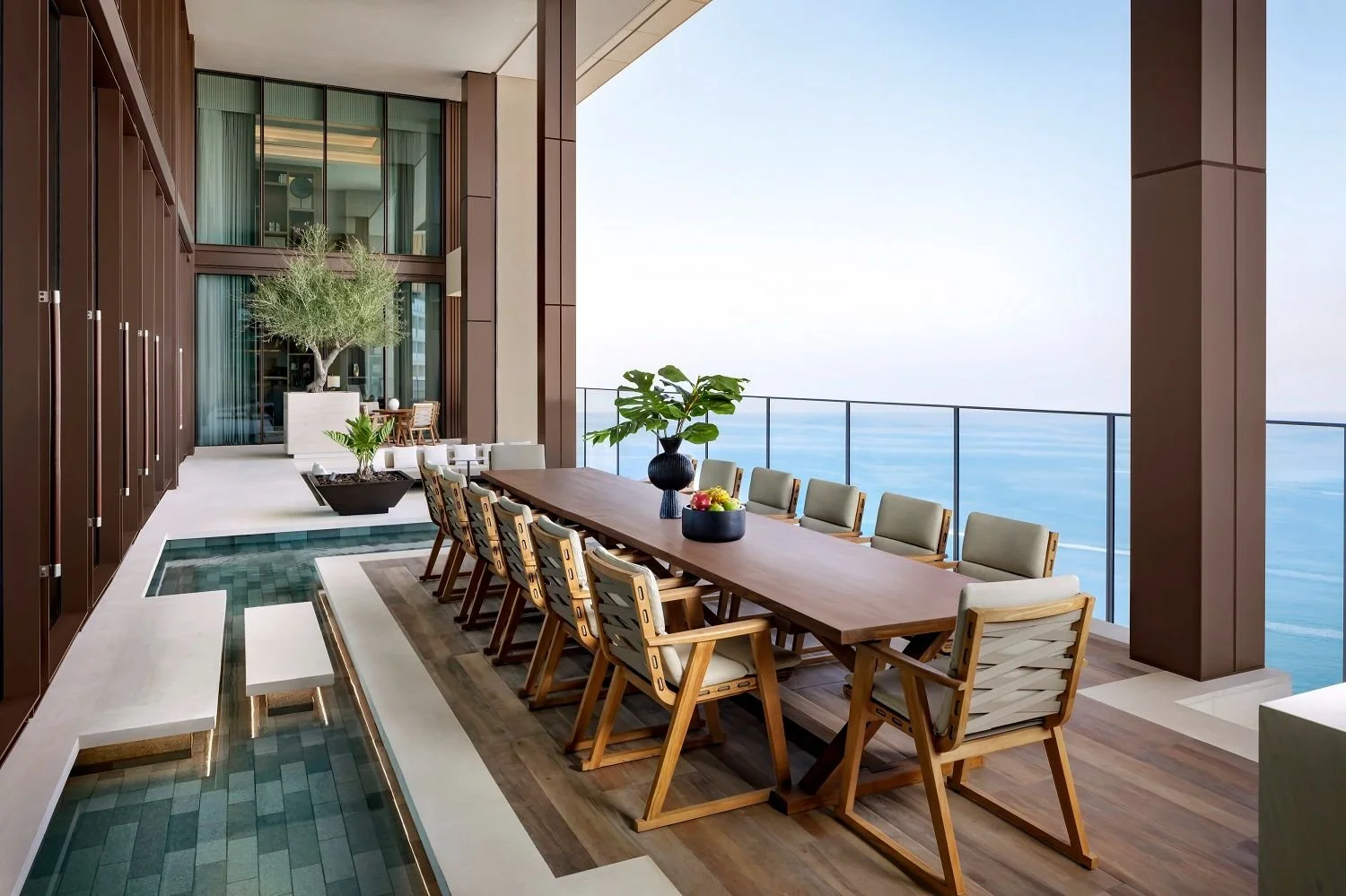 Elegant outdoor balcony with a long wooden dining table, cushioned chairs, decorative plants, and ocean view.
