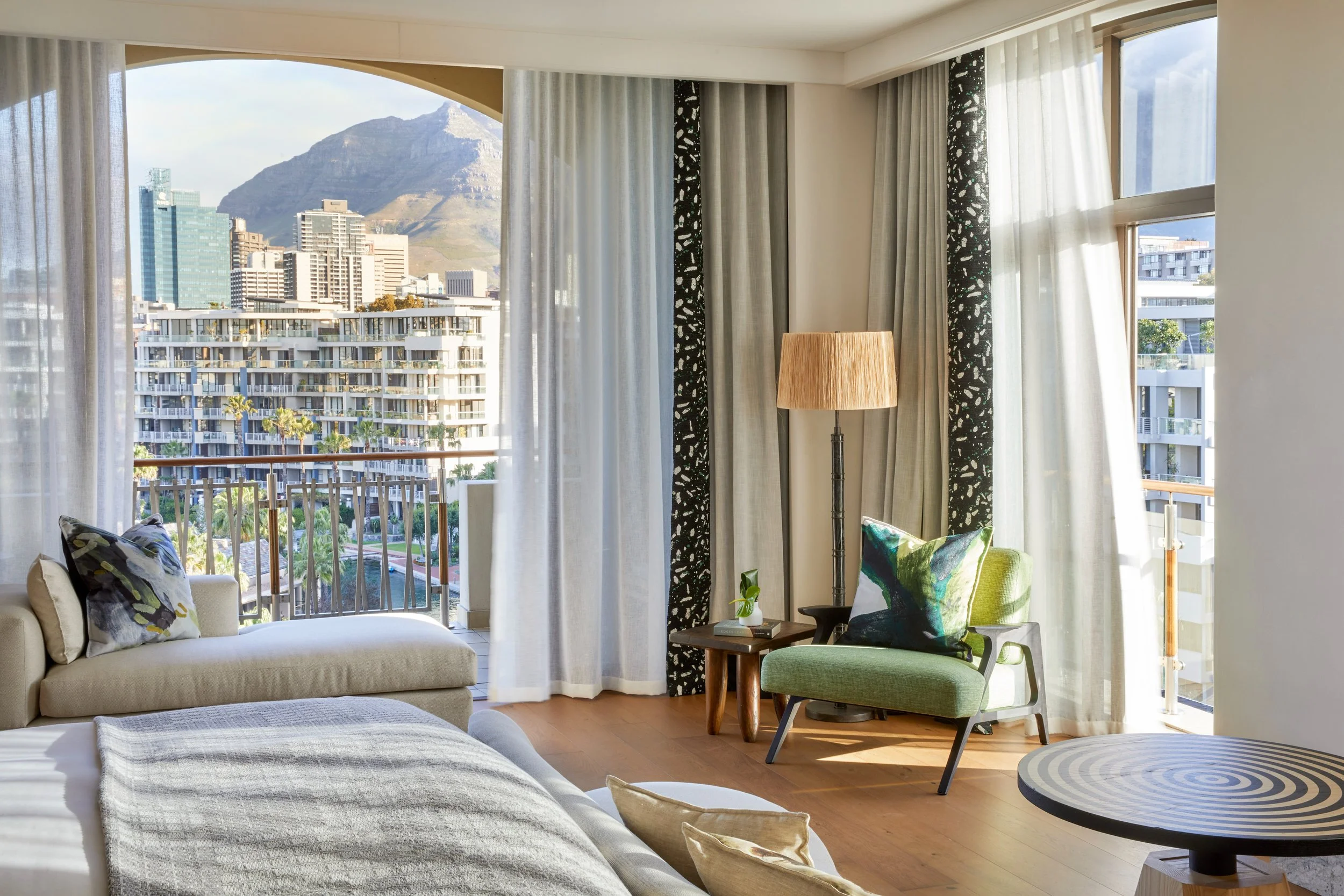 Living room with large windows showing cityscape and mountains, white sofa with patterned pillows, green chair, wooden side table with plant, and a round black and white patterned table.