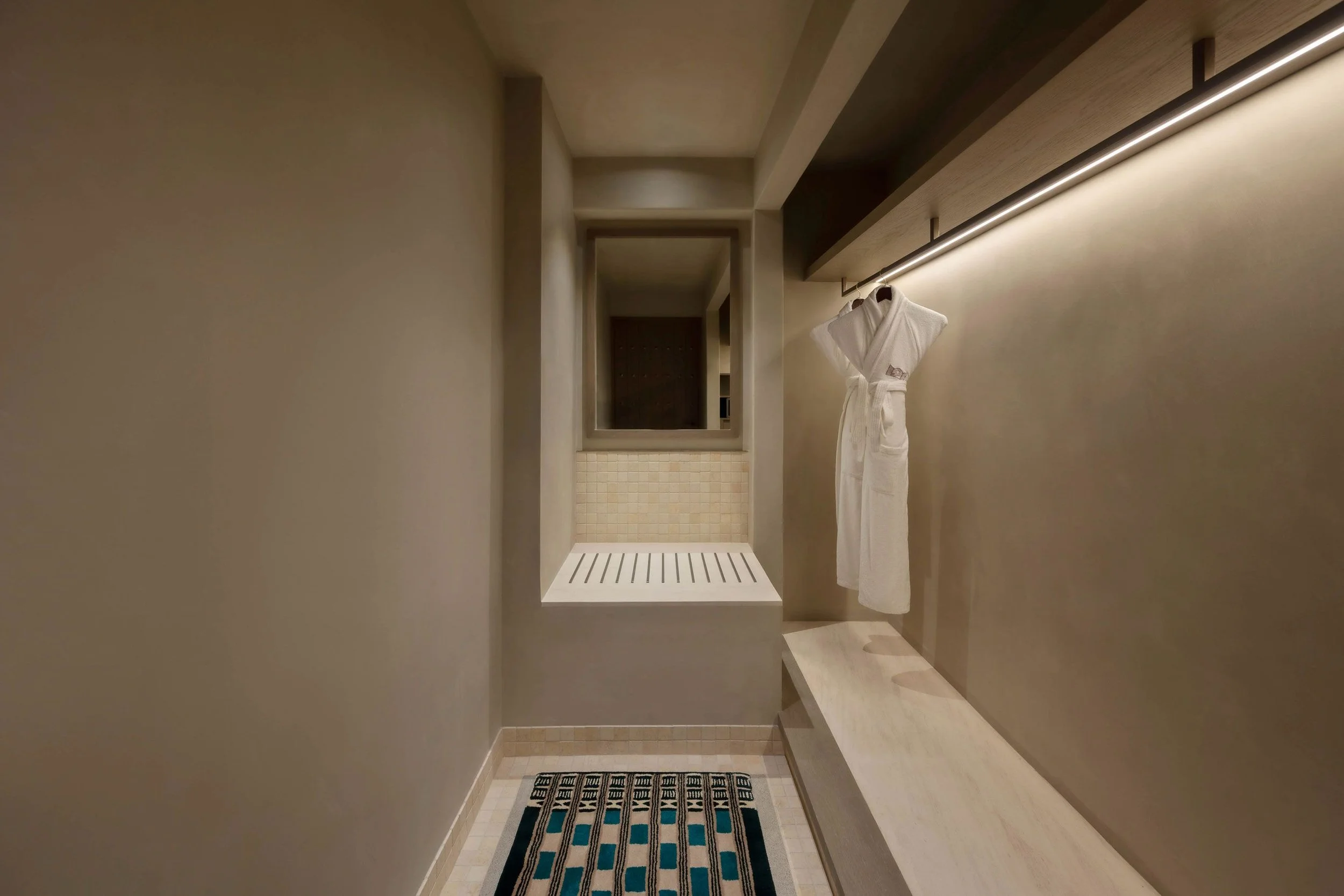 Modern walk-in shower with beige walls, a built-in bench, a towel hanging on a rail, and a patterned rug on the floor.